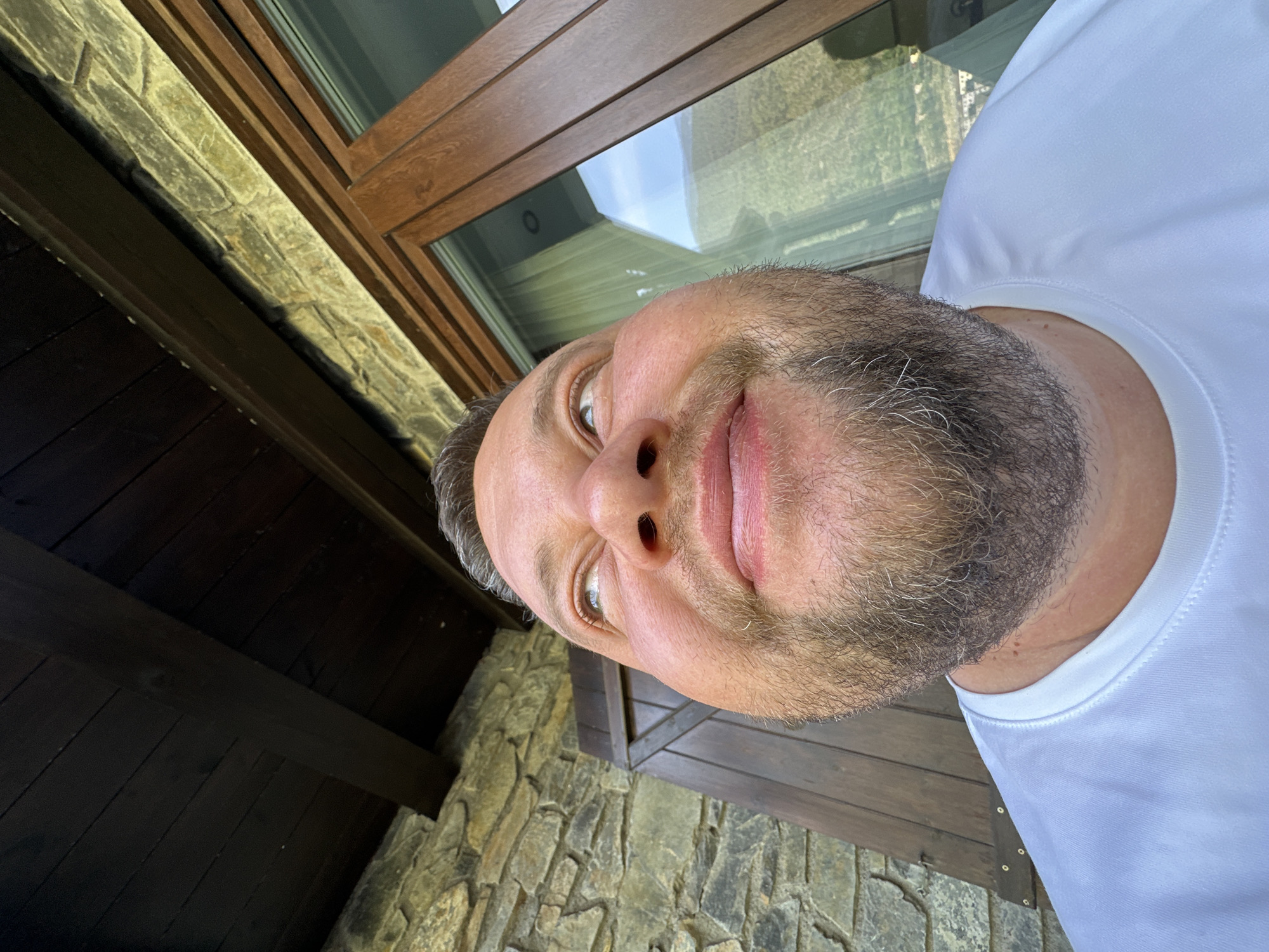 A close-up, slightly low-angle portrait depicts a middle-aged man with a beard, wearing a white t-shirt. His face is positioned centrally in the frame, with a slight smile. The background reveals architectural details, including a brown window frame with a visible reflection and a stone wall. Dark wooden elements are also present. The location is Encamp, Andorra, evidenced by the distinctive architecture and likely geographic context. The man is the primary subject, and the architectural elements suggest an indoor or partially enclosed space.