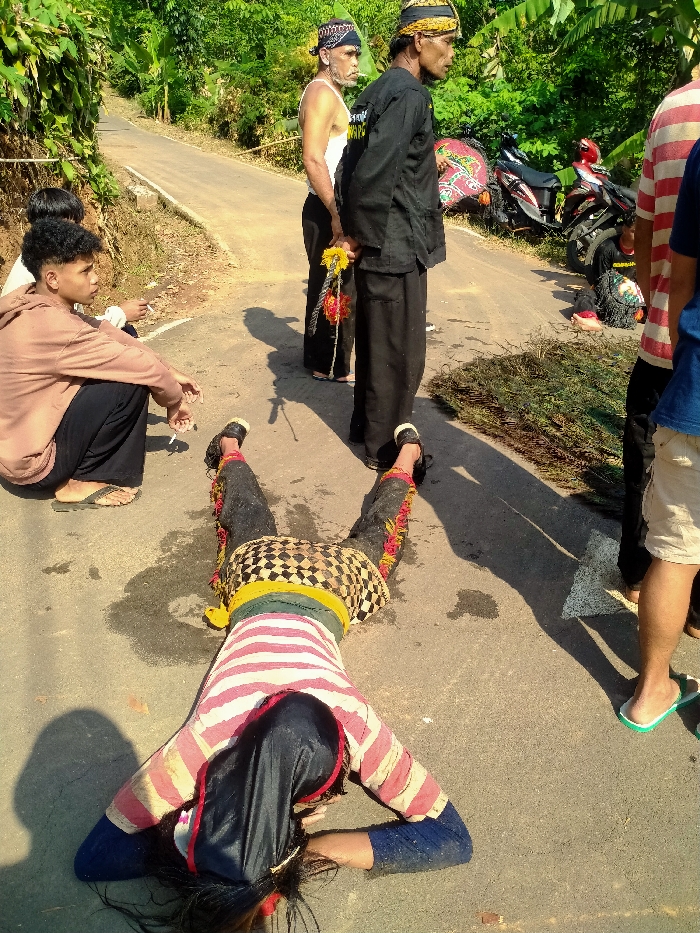 An outdoor scene in Gempolan, Indonesia, depicts a person lying prone and face down on an asphalt road. This individual wears a red and white horizontally striped long-sleeved shirt over a blue undershirt, a black head covering partially revealing dark hair, and traditional lower garments consisting of a patterned fabric with a yellow and green waist band, covering black pants adorned with red and yellow fringe. Their arms are bent, with hands positioned near the head.
Several other individuals are present. To the mid-right, a man in a black long-sleeved shirt and a dark head covering stands, holding a decorative yellow and red bundled object in his left hand. Behind him, another man in a white tank top and a dark head covering stands, smoking a cigarette. To the mid-left, a young man with dark curly hair, wearing a light brown hoodie and dark pants, sits on the asphalt, smoking. A person in a blue shirt and light shorts is partially visible to the far right.
Two motorbikes, one red and black, the other black, are parked off the right side of the road. A colorful, patterned cloth is draped over the seat of the red and black motorbike. A pile of green foliage is situated on the asphalt to the right of the prone figure. The asphalt road, which shows some damp patches, ascends into the background, flanked by dense green vegetation. The scene is brightly lit by direct sunlight, casting distinct shadows.