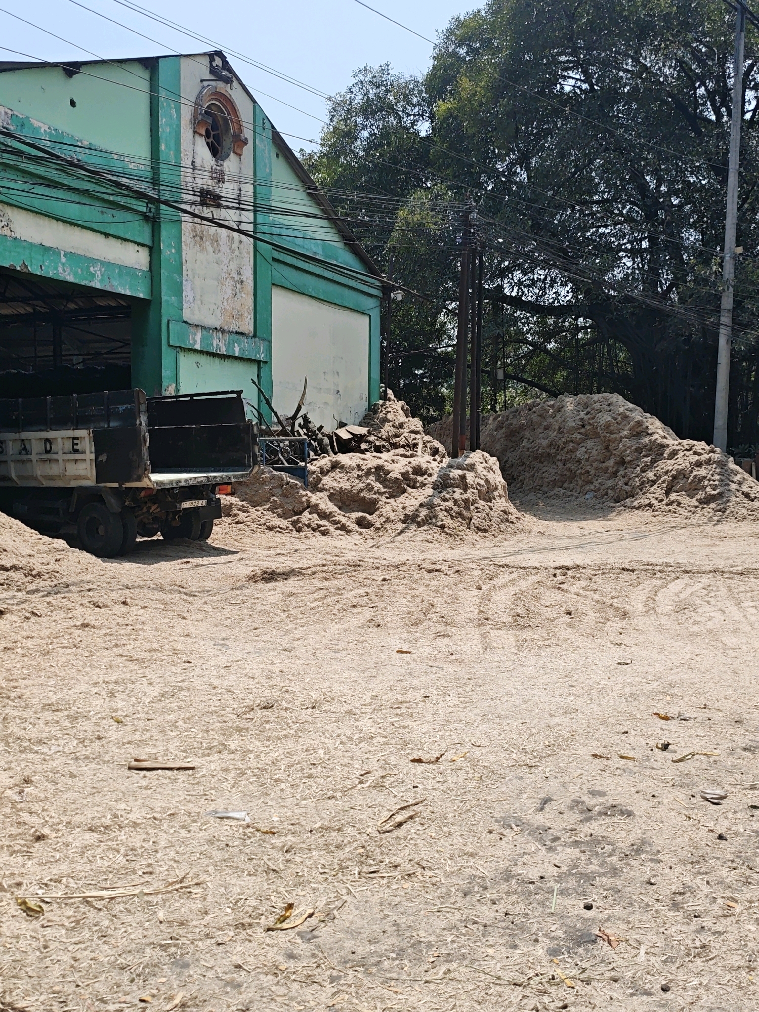 The image depicts an outdoor scene with a large pile of shredded material, possibly wood chips or sawdust, dominating the foreground and midground. A light-colored truck with a dark bed is parked in the left-midground, partially obscured by the material. The truck has the word "STADE" visible on its side. In the background, a weathered building with peeling green and white paint is visible. The building has a circular, decorative window opening near the apex of its gable. Trees with dense foliage are present behind the building and to the right. Electrical wires crisscross the upper portion of the image, supported by utility poles. The ground is covered with the same shredded material. The scene appears to be an industrial or agricultural area, likely in Kediri City, Indonesia, given the context. There are no people clearly visible in the image.