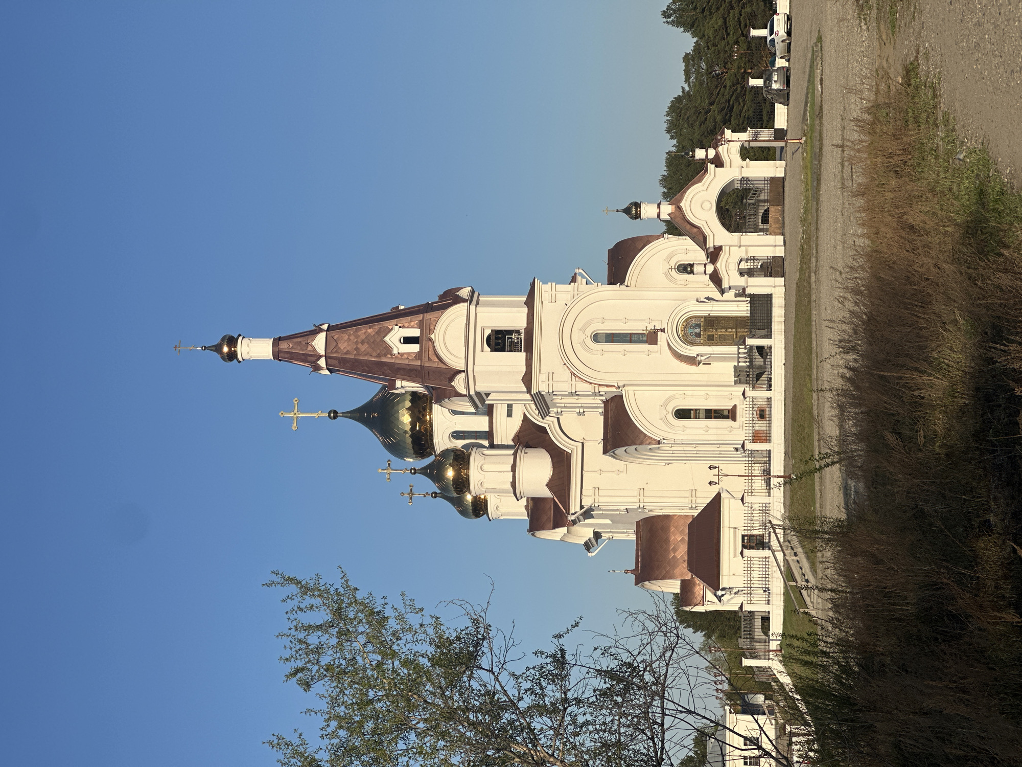 The image depicts a white Orthodox church with copper roofing and golden domes in Shelekhov, Russia. The church stands under a clear blue sky. A gravel path borders the building on one side, with vegetation including grass and trees. A fence and possibly a parking area with vehicles are visible in the background. A bare tree branch is in the foreground. The image appears to be taken at an angle.