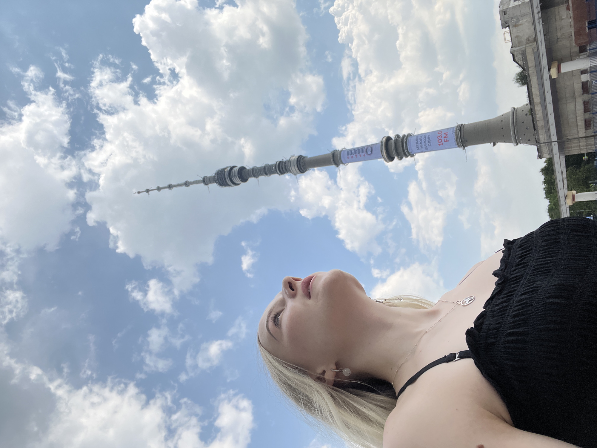In Khimki, Russia, a young Caucasian woman with blonde hair is captured from a low angle, looking upwards at a cloudy sky and a tall television tower. She is wearing a black, ruffled, strappy top and jewelry, including earrings and a necklace with a circular pendant. The television tower, a prominent structure in the city, dominates the background. It is a cylindrical tower with multiple levels and is adorned with banners displaying text and "103.0 FM". The sky is a mix of blue and white clouds, suggesting a bright but partly overcast day. In the lower right portion of the image, a section of a concrete structure, possibly part of the tower's base or an adjacent building, is visible.