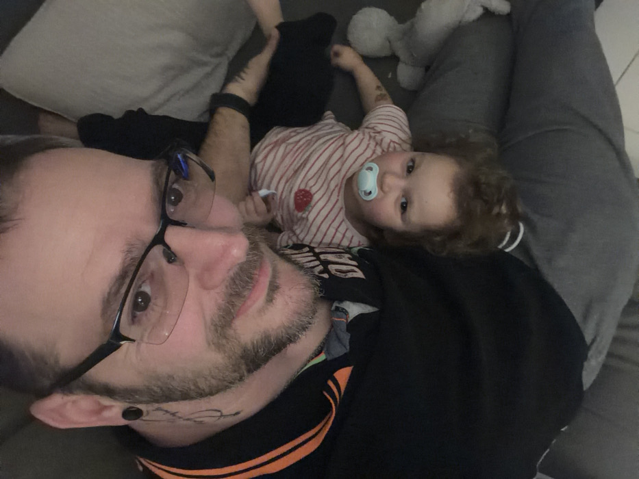 The image displays an adult male and a young child in an indoor setting, potentially within the city of Trassem, Germany, although specific visual identifiers for the precise location are not present. The adult male occupies the upper portion of the frame, facing the camera with a slight smile. He has a short beard and mustache, wears black-framed glasses, and a black polo shirt with orange stripes on the collar. A black ear tunnel is visible in his left earlobe, and a tattoo, appearing as script or abstract lines, extends from his left neck downwards. A black fitness tracker is present on his right wrist. Below him, oriented with its head towards the bottom of the frame, is a young child with light brown, curly hair. The child has a light blue pacifier in its mouth and wears a light-colored shirt with thin red horizontal stripes and a small red strawberry appliqué on the chest. A dark mark or small tattoo is visible on the child's left arm. The child's legs are covered by grey pants. A light-colored stuffed animal is partially visible on the right side of the frame, near the child's legs. The immediate background consists of blurred fabric, consistent with furniture upholstery.