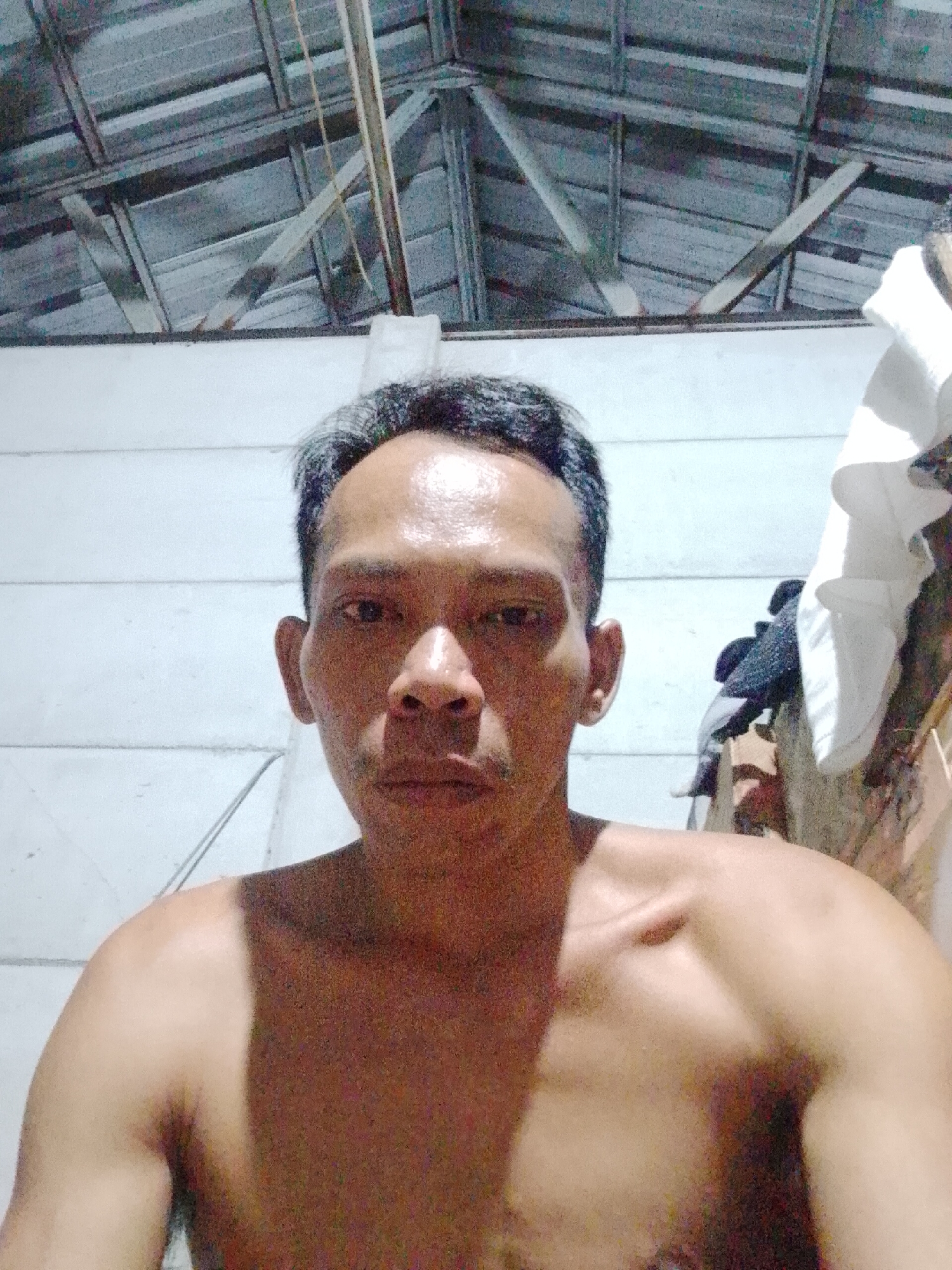 A shirtless middle-aged man with short dark hair and graying temples is facing the camera indoors. He is lit from the front, with a plain white wall behind him. Above the wall is a corrugated metal roof structure with wooden supports. To the right, a hanging white cloth and dark clothing items are visible. The location is in the general area of Bukit Ketok, Indonesia based on source information.