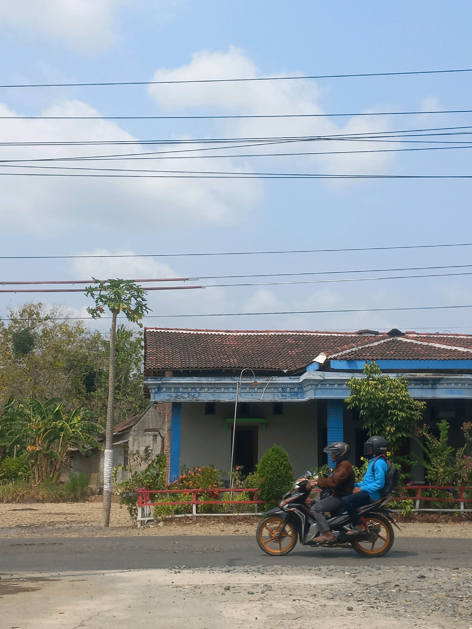 Two individuals are riding on a motorcycle on a paved road. The person in the front is wearing a brown jacket and a helmet, while the person in the back is wearing a blue shirt and a helmet with a backpack. The motorcycle is black and orange with orange rims. They are traveling past a residential building with a red-tiled roof and blue trim. In the background, there are trees and vegetation, including what appears to be a papaya tree. Power lines are visible in the sky above. The scene appears to be in a rural or semi-rural area in Baturetno, Indonesia.