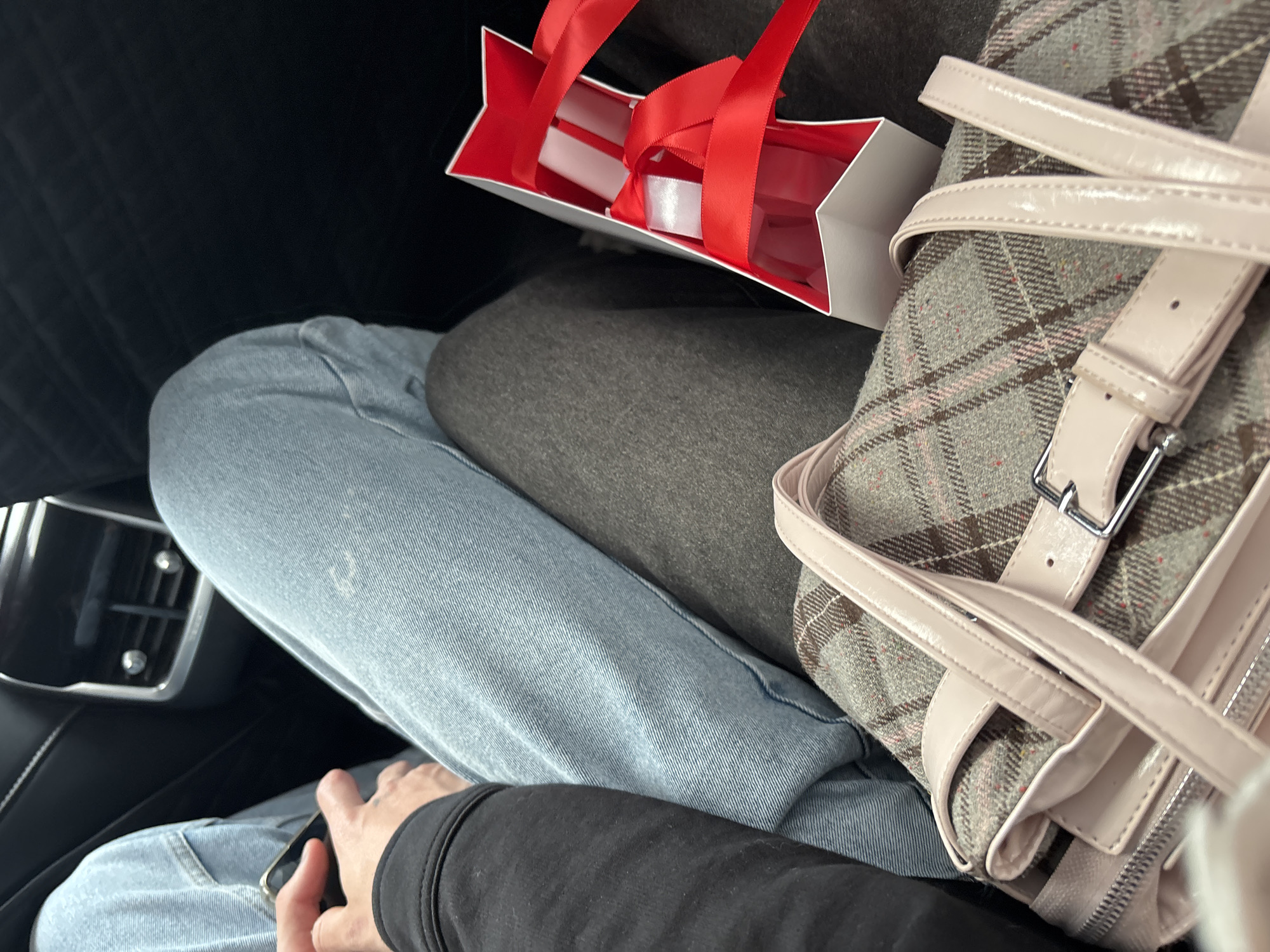 A close-up, eye-level shot shows the lower bodies of two people seated next to each other, possibly in the back of a vehicle. The person on the left wears a black jacket and light blue jeans, holding a black smartphone. The person on the right wears light blue jeans and grey pants. In the foreground, a plaid tote bag with pink leather straps rests on the lap of the person on the right. To the right of the bag, a white gift box with red ribbon is visible. The background includes a portion of a car's interior with black quilted upholstery. The scene is captured indoors.