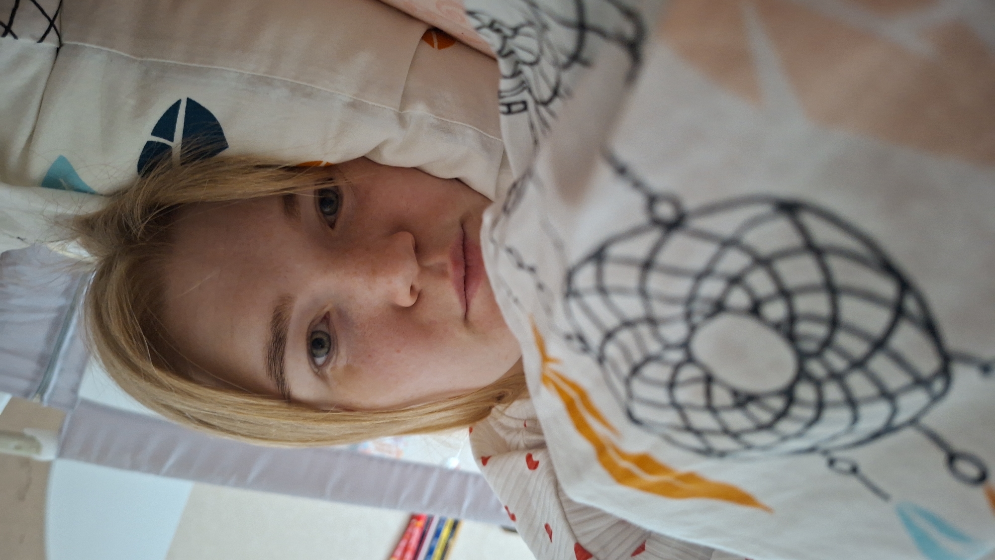 The image shows a person with fair skin and blonde hair lying down, possibly in bed, under patterned bedding. The patterns include a dreamcatcher design and geometric shapes in colors such as blue and orange. The person's face is visible, displaying freckles and neutral expression. In the lower-left corner, a glimpse of what appears to be a children's playpen or crib, and colored pencils or markers, can be seen. The image was taken in an indoor setting, likely a bedroom, but the specific location of Chaikovsky, Russia, cannot be determined from the provided visual information.