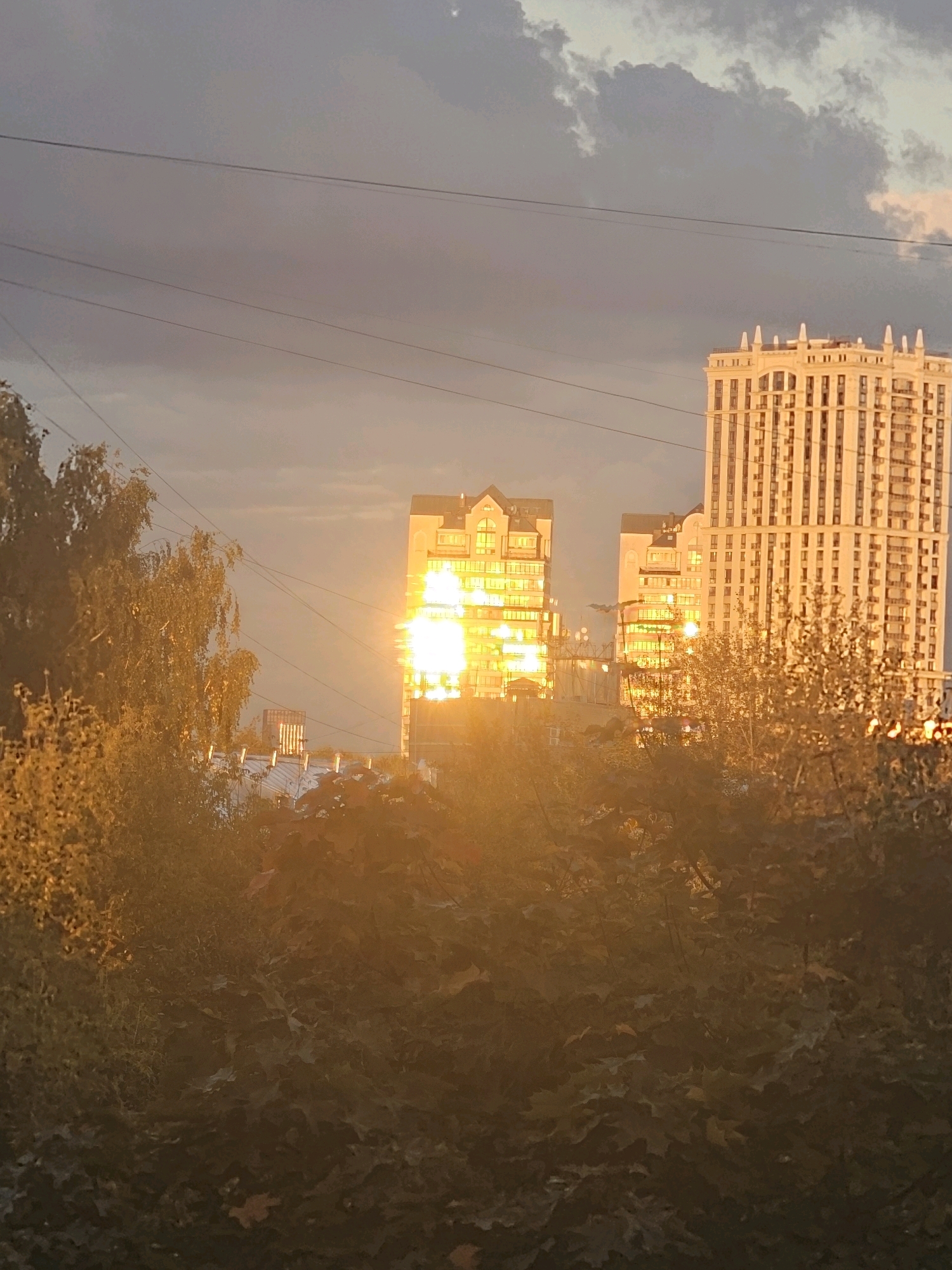The image captures an outdoor scene in the city of Khimki, Russia, during what appears to be sunset or sunrise. The sky is filled with clouds, with a mix of grey and hints of pink or orange, suggesting transitional lighting conditions. Several power lines stretch horizontally across the frame, obscuring parts of the sky.
In the background, two prominent apartment buildings are visible. The building on the right is tall and slender, with a facade characterized by numerous vertical lines and a decorative top. The building on the left is shorter and wider, also featuring a multi-story design with visible windows. Both buildings are illuminated by the setting or rising sun, which reflects intensely off their glass windows, creating bright, almost blinding streaks of light.
In the foreground and midground, there are trees and foliage. The leaves on the trees are a mix of green and orange or yellow hues, indicating an autumn or late summer season. The trees are dense, with some branches and leaves partially obscuring the view of the buildings. The lighting from the sun casts a warm, golden glow over the entire scene, particularly on the trees and the lower parts of the buildings. The overall impression is one of urban landscape bathed in the dramatic light of dawn or dusk. No people are visible in the image.
