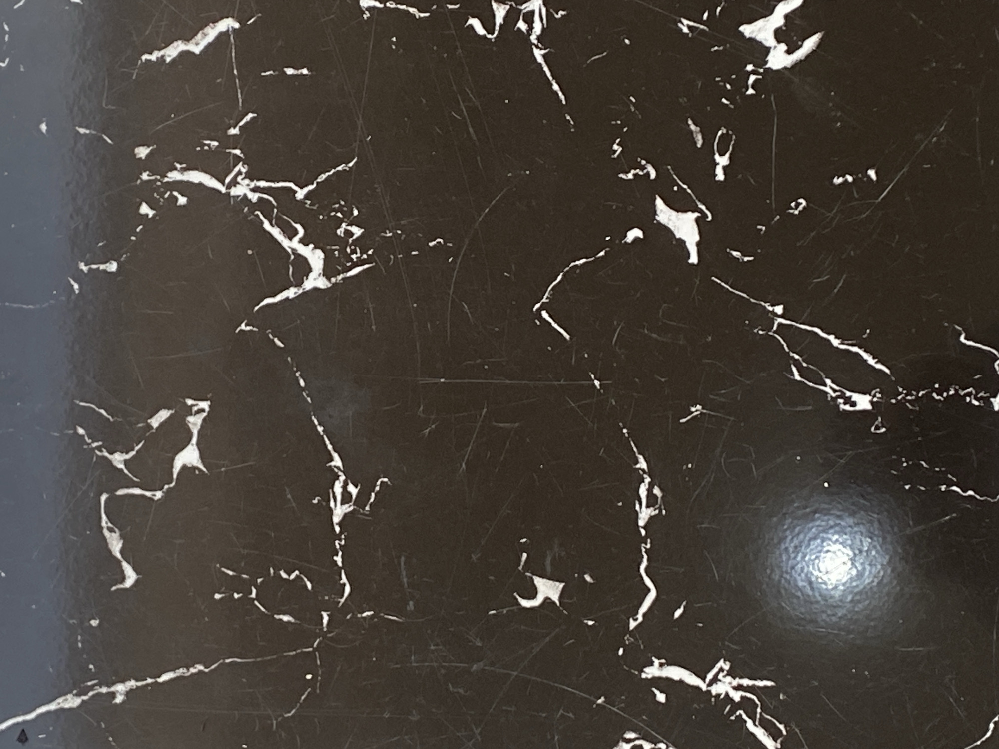 The image is a close-up, top-down view of a dark, possibly black, marble-like surface. The surface is adorned with irregular white veins, characteristic of marble. There are numerous fine scratches visible across the surface, suggesting wear or use. In the lower left quadrant of the image, a bright, circular reflection is present, likely from a light source. No elements suggestive of a specific location in Jauro Yinu, Nigeria, are discernible.
