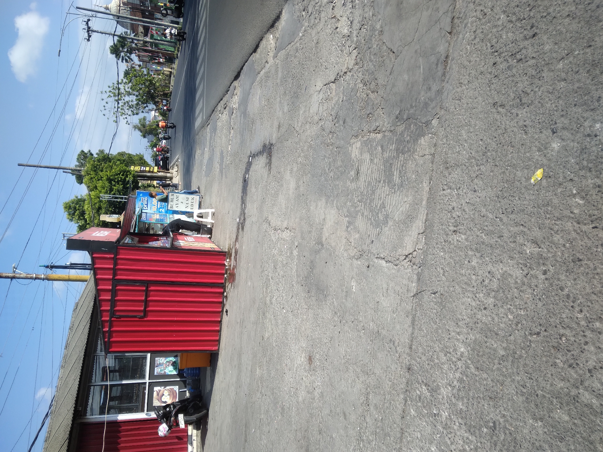 A daytime, eye-level, outdoor shot captures a street scene in Purwokerto Kidul, Indonesia. The foreground is dominated by a cracked, grey concrete sidewalk with patches of darker, wet pavement and scattered debris. To the left, a red corrugated metal kiosk with a black frame stands adjacent to a red building with a sloped grey roof and large windows. A black motorcycle with a white helmet is parked to the left of the kiosk. Behind the kiosk, a person in dark clothing is visible. Further down the sidewalk, a white plastic stool and a blue sign are present. The street, a mix of asphalt and zebra crossing markings, stretches into the distance, populated by several motorcycles and cars. Trees and utility poles line the right side of the street, with buildings and a dome-shaped structure visible in the background. The sky is blue with scattered white clouds.