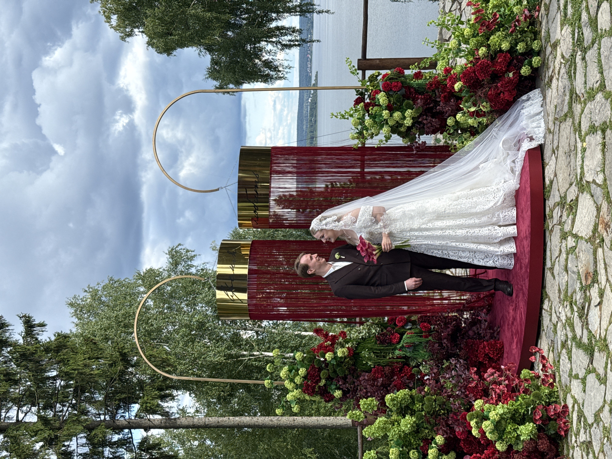 A wedding ceremony is taking place outdoors in Kanabekovo, Russia, near a large body of water. A newlywed couple stands on a circular red carpeted platform. The groom wears a dark brown suit and the bride is in a white lace wedding gown and veil. The couple is looking at each other and smiling. They are surrounded by elaborate floral arrangements in shades of red and green, and two tall, cylindrical structures draped with red fringe and topped with gold. Behind them, a calm body of water is visible, bordered by trees and a cloudy sky. The ground in front of the platform is paved with large, irregularly shaped stones.
