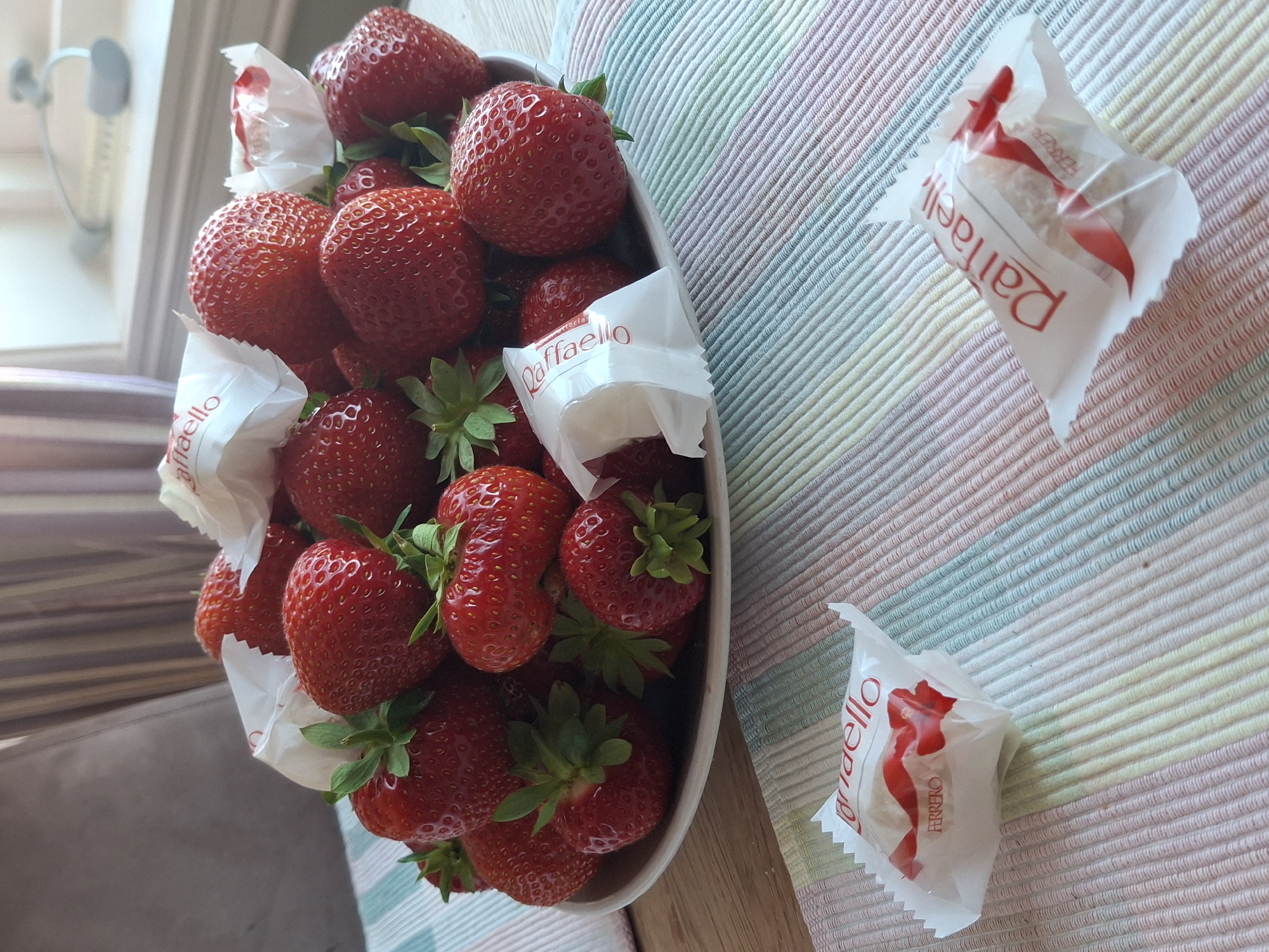 This media file depicts a bowl of fresh strawberries, artfully arranged on a striped placemat. The strawberries are a vibrant red, with their green leaves still attached, suggesting they are ripe and recently picked. Interspersed among the strawberries are individually wrapped Raffaello chocolates, their white paper wrappers featuring the distinctive red logo. Two of these chocolates are also placed on the placemat, separate from the bowl, showcasing the packaging and the chocolate itself, which appears to be white and round.
The setting suggests a casual, possibly domestic environment. The placemat, with its pastel stripes, adds a touch of color and texture to the scene. The lighting in the image is soft and natural, hinting at daytime. In the background, out of focus, there are glimpses of what appears to be an indoor setting with a window and possibly some furniture, though these details are not clearly discernible. The overall impression is one of a sweet treat being offered or enjoyed, evoking a sense of simple pleasure. The visible text on the wrappers clearly reads "Raffaello" and "FERRERO".