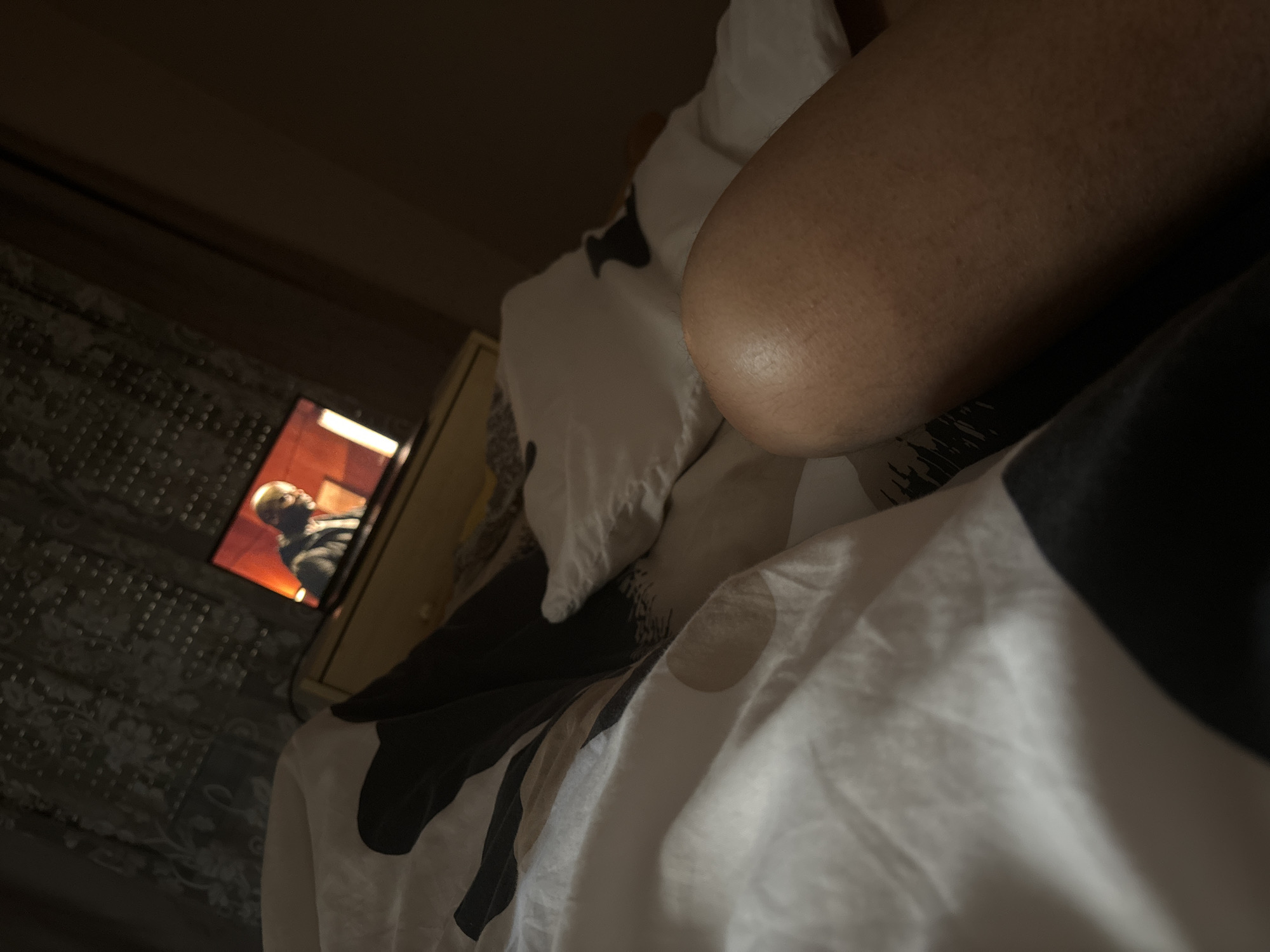 The scene is indoors, likely a bedroom. A person is lying in bed, with a raised knee prominently in the foreground. Bedding includes a white pillow and a patterned duvet or blanket with dark and light sections. A television is visible in the background, displaying a man. The room is dimly lit. The location, Budva, Montenegro, cannot be confirmed from the provided visual information.