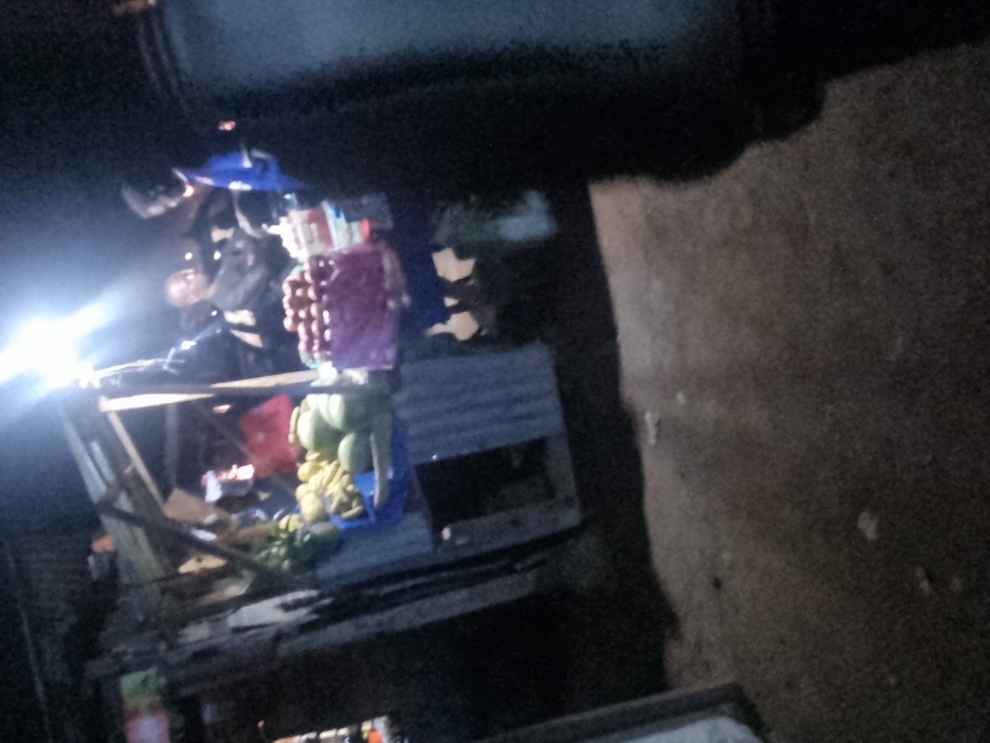 The image depicts a nighttime scene at a market stall, likely in Jos, Nigeria. A bright light illuminates the stall, where various fruits, including bananas and what appear to be apples or tomatoes, are displayed in baskets. Two individuals are visible at the stall. One, wearing a blue shirt, appears to be interacting with another person who is facing away from the camera, partially obscured by shadow. In the background, another person in a red top is also visible. The stall itself is constructed of makeshift materials, including corrugated metal and wood. The ground appears to be dirt or gravel. The overall atmosphere suggests a busy, open-air market operating after dark.