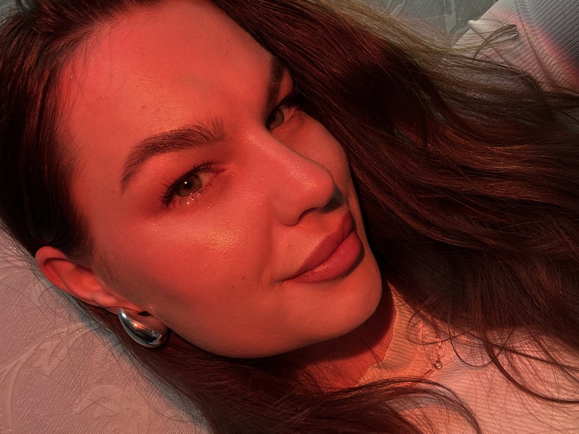 A close-up portrait features a young adult female reclining, with her head tilted, looking towards the viewer. Her long, wavy, dark brown hair cascades around her and onto the textured background. The entire scene is bathed in a distinct red-orange light, creating a warm and dramatic ambiance.
Her skin appears smooth and radiant, highlighted by the strong red illumination on her forehead, cheekbones, and nose. She has light-colored eyes, which appear greenish-grey under the lighting, with visible eyelashes and well-defined eyebrows. A subtle sparkle or glitter is visible near the inner corner of her left eye. Her full lips are adorned with a reddish-brown or plum lipstick, and her expression conveys calmness with a slight, enigmatic smile.
She wears a large, smooth, silver-toned, curved earring in her visible left earlobe. A delicate, thin chain necklace with small rings is partially visible against the neckline of her light-colored, subtly textured top, possibly a white high-neck garment. The background consists of a soft, light-colored fabric with a patterned surface, likely a pillow or bedding. The intense red lighting suggests an indoor setting, possibly evening or created with artificial mood lighting. No text is visible in the image. The location is Chaikovsky, Russia.