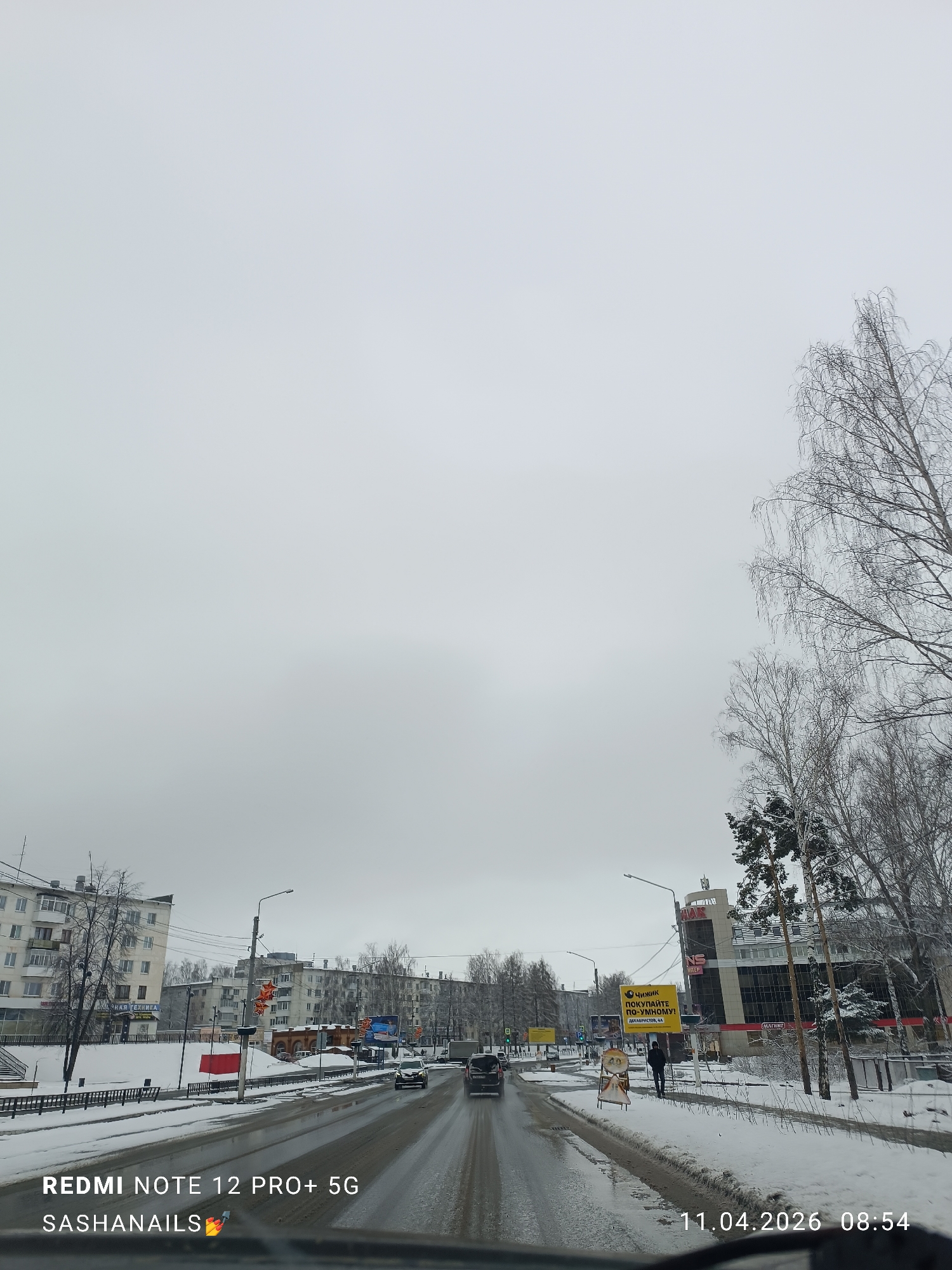 The image depicts a street scene in the city of Chaikovsky, Russia, on April 11, 2026, at 08:54. The view is captured from within a vehicle. The weather appears to be overcast, and snow covers the ground on either side of the street. The street surface is wet, indicating recent snowfall or melting. Several cars are traveling in both directions on the road. On the right side of the street, a pedestrian walks on the snowy sidewalk. A large advertisement featuring the words "Chizhik BUY SMARTLY!" is visible near a multi-story building with a "Magnet" sign. The residential buildings line the left side of the road. Trees with bare branches stand on both sides of the street, and the overcast sky dominates the upper portion of the frame. The camera is a Redmi Note 12 Pro+ 5G, and the watermark "SASHANAILS" is also present.