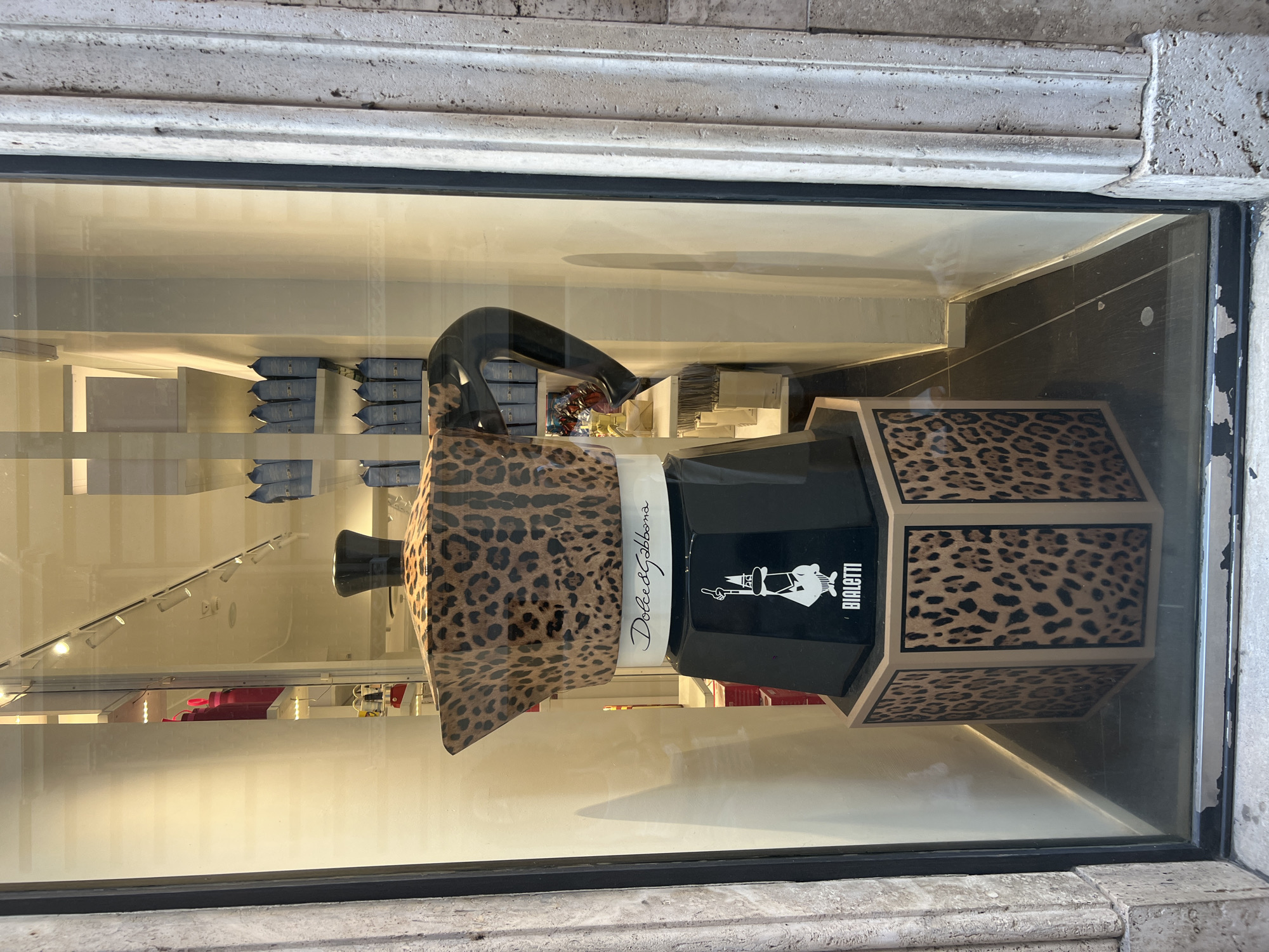 This is a photograph taken in Rome, Italy, in a shop window. The focal point is a large, decorative Moka pot, a type of stovetop espresso maker. The Moka pot is covered in a leopard print pattern, and the word "Dolce&Gabbana" is written in cursive on a white band around its middle. Below the leopard print section, the pot is black and features the Bialetti logo, which is a silhouette of a man with a mustache, raising his hand. The Bialetti logo is also printed on the black hexagonal base of the Moka pot. The base itself is covered in leopard print panels.
Behind the Moka pot, inside the shop, shelves are visible with various items. On the left, a bright pink object, possibly a bag or clothing item, is displayed. Further back, rows of blue packaging, likely coffee bags, are neatly arranged on shelves. The shop interior is brightly lit with overhead lights.
The shop window is framed by a stone pillar on the right side of the image, which has a rough, textured surface and vertical fluting. The glass of the window reflects the surroundings, including a blurry image of what appears to be another building or street scene. The bottom edge of the window shows some peeling paint or wear. The overall scene suggests a retail display, likely in a fashion or home goods store.