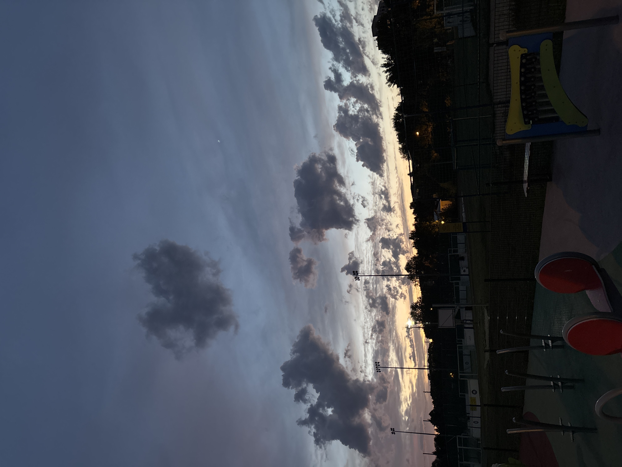 The image depicts a playground scene at dusk, likely in Lublin, Poland, given the context. The sky is a deep, dusky blue with streaks of orange and yellow from the setting sun. Dark, dramatic clouds are scattered across the sky, with one cloud in the upper left quadrant resembling a heart shape. In the distance, there are sports fields with bright floodlights, trees, and the silhouette of buildings. In the foreground, parts of playground equipment are visible, including red circular elements and a musical xylophone-like structure. The overall atmosphere is serene and transitioning from day to night.