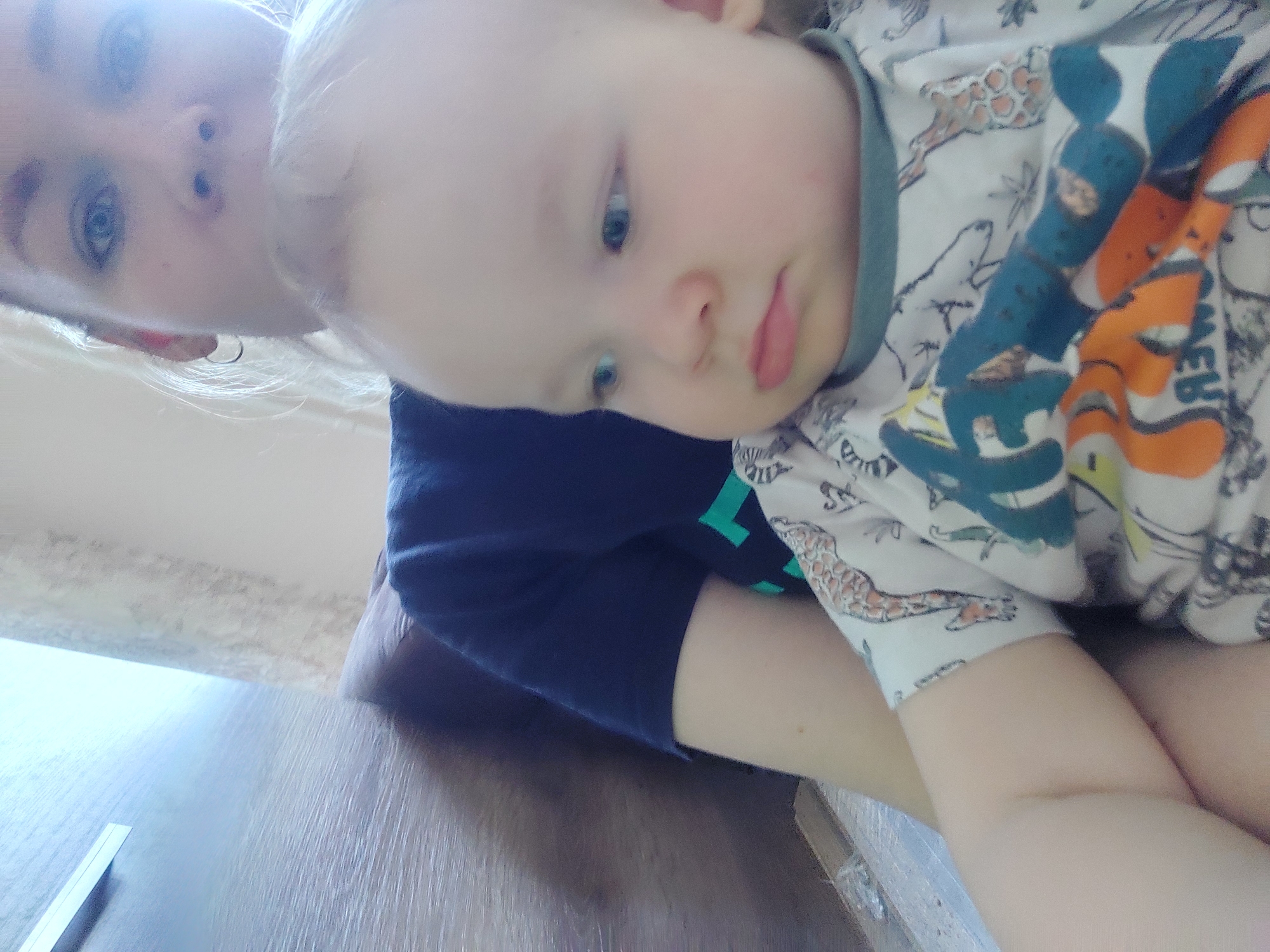 A close-up, tilted shot shows a fair-skinned child with blue eyes in the arms of an adult. The adult, also fair-skinned with blue eyes, is positioned in the upper left corner of the frame. The child is wearing a white, patterned shirt with images of animals and blue and orange shapes, while the adult wears a dark blue shirt. The location is Chaikovsky, Russia. The foreground shows a wooden surface.