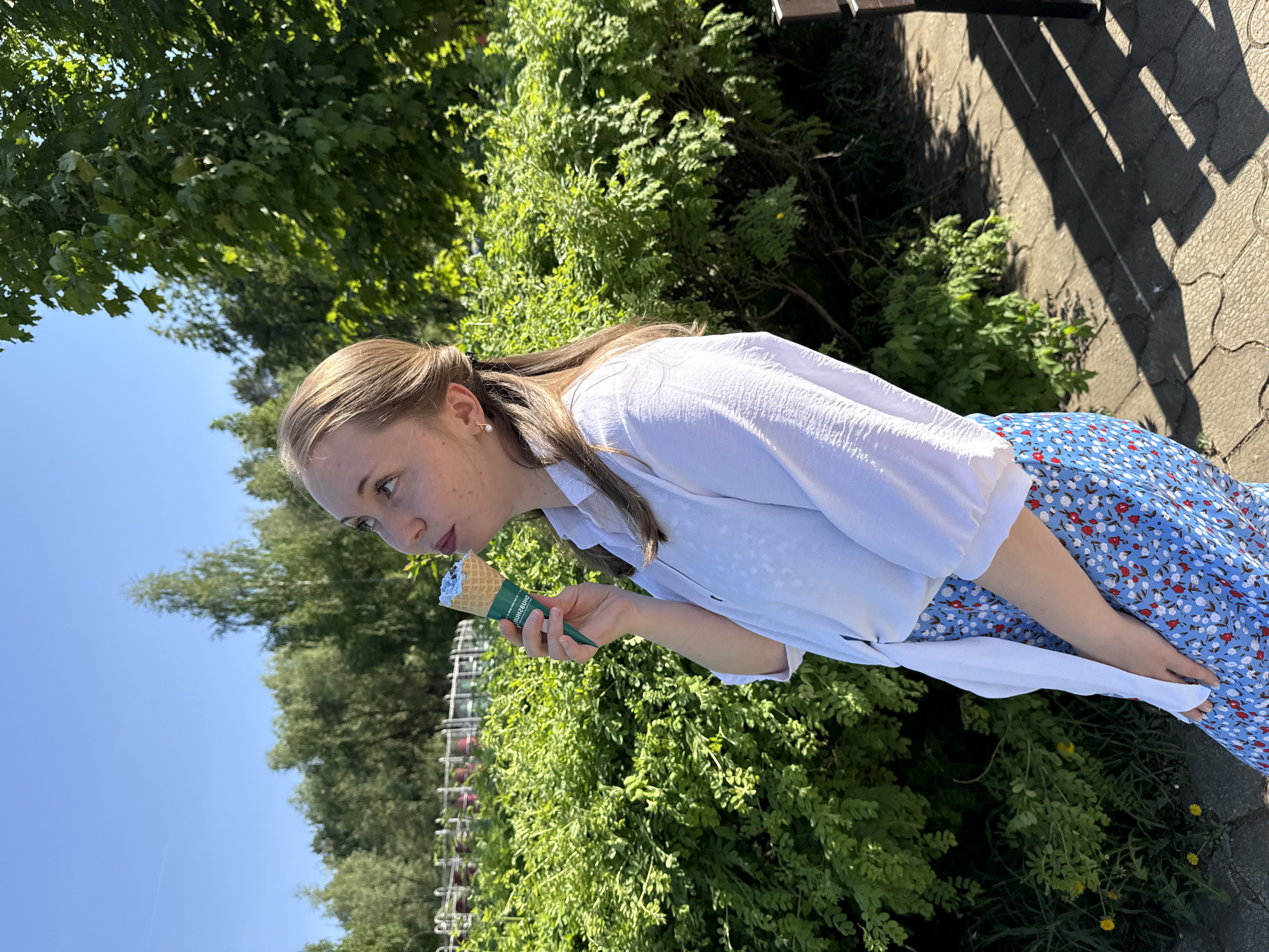 A young woman stands outdoors with greenery and a clear blue sky in the background, holding an ice cream cone. She is wearing a white, button-down shirt over a blue floral skirt. The location is likely a park or garden in an urban setting.