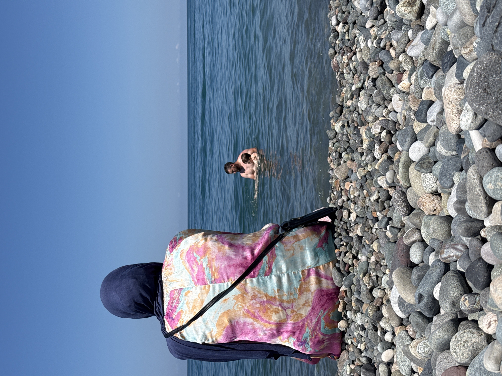 A person wearing a dark blue hijab and a colorful pink, blue, and orange patterned top sits on a pebbled beach facing away from the camera, looking out at the ocean. The person has a dark bag slung across their body. In the water, a man is holding a small child. The sea is blue with gentle waves, and the sky is clear and bright blue. The foreground is filled with smooth, grey and colored pebbles of various sizes.