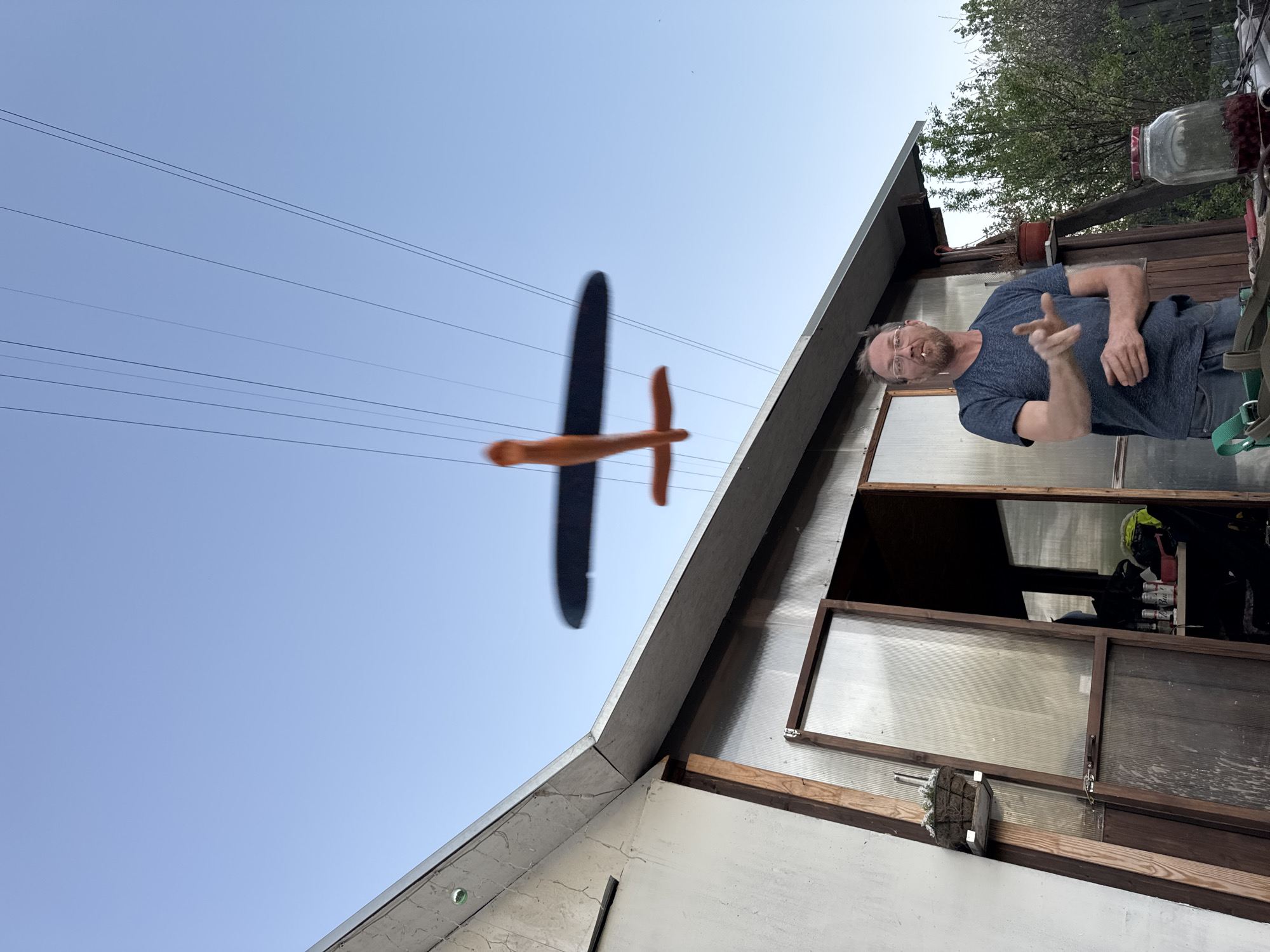The image shows a man standing next to a building with an open window. The man is wearing a gray t-shirt and jeans, and he is pointing at the camera. A toy airplane is suspended from wires in front of the building. The location is Chaikovsky, Russia.