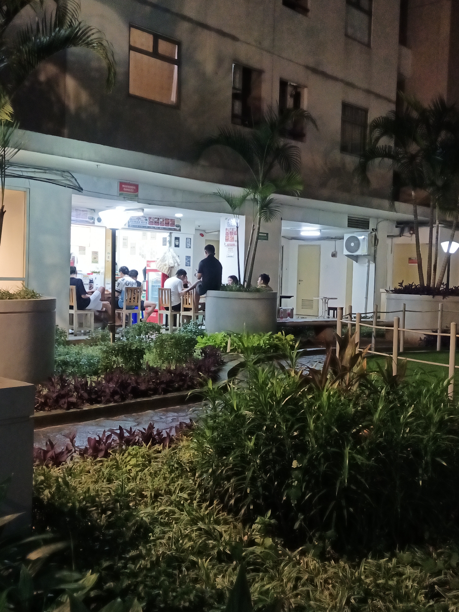 The image depicts a dimly lit outdoor seating area of a commercial establishment, possibly a restaurant or cafe, in South Jakarta, Indonesia. The establishment is situated on the ground floor of a building with multiple windows visible on the upper floors. Several patrons are seated at tables, some indoors and some outdoors. One person, wearing a black t-shirt, is standing near an open doorway, facing away from the camera. Lush green foliage and planters surround the seating area, and a low wooden fence is visible in the background. The lighting suggests it is nighttime. The overall impression is one of a casual dining or gathering place.