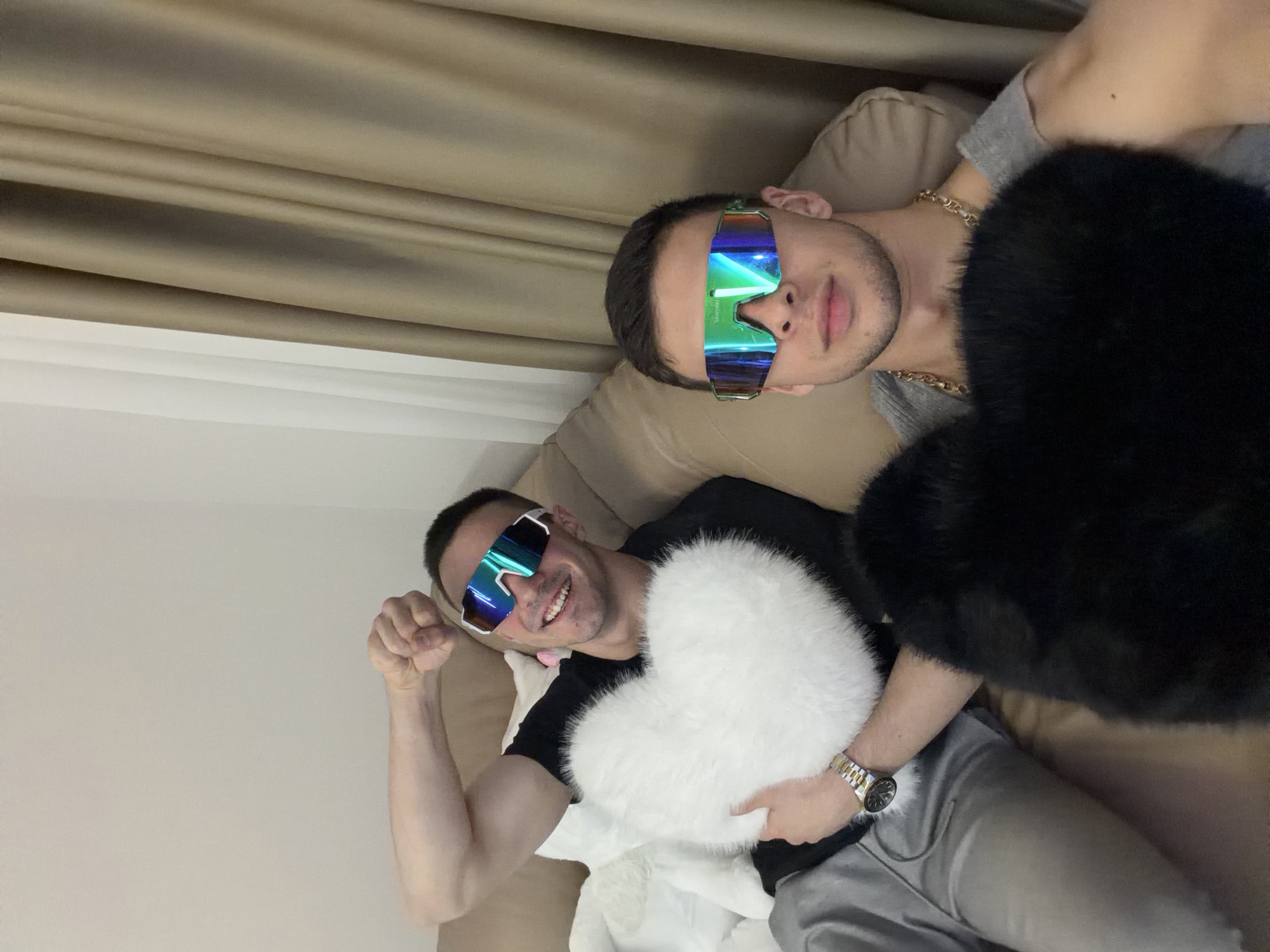 Two men wearing reflective sunglasses are posing on a tan couch. One man, wearing a black shirt and grey sweatpants, is holding a white heart-shaped pillow while flexing his arm. The other man, wearing a grey shirt and a gold chain, is partially obscured by a large black fur-like object. The background includes a tan curtain on the upper left and a light-colored wall on the lower left. Location: Unknown, Unknown.