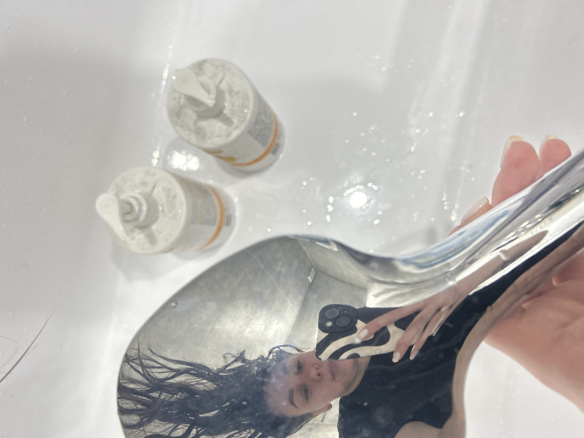 The image displays an indoor scene, consistent with a bathroom or wash area, characterized by white, wet surfaces. The specific location within Chaikovsky, Russia, is not ascertainable from the visual evidence.
In the foreground, a female individual's hand holds a curved, chrome-plated object, likely a shower head or faucet component. The object's reflective surface captures a selfie. The reflection reveals the individual's face, dark, wet, and disheveled hair appearing to float upwards, and a dark-colored top. Her right hand, visible in the reflection, holds a black smartphone with a white and black patterned case that features a triple-lens camera array. Her fingernails are painted a light, possibly white or off-white, color.
In the background, on a wet, white surface, two identical white pump-dispenser bottles, each with an orange band or label near its base, are visible. Scattered water droplets are present across all visible surfaces, including the reflective object and the white background. A few strands of dark hair are observed on the white surface above the reflective object. A grey tiled wall is partially reflected in the chrome object.