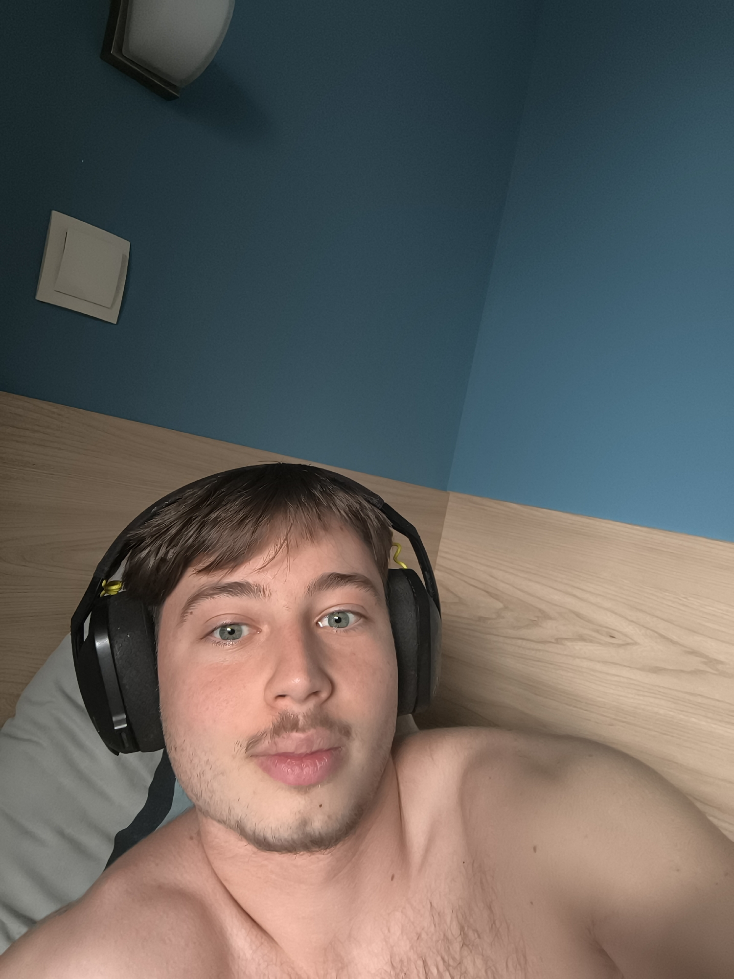 A fair-skinned young man is lying down, wearing black headphones with neon yellow accents. He has light brown hair, blue eyes, and stubble. He is shirtless. The background consists of a light wood headboard and a blue wall with a white light switch and a modern light fixture. The location cannot be determined based on the image.