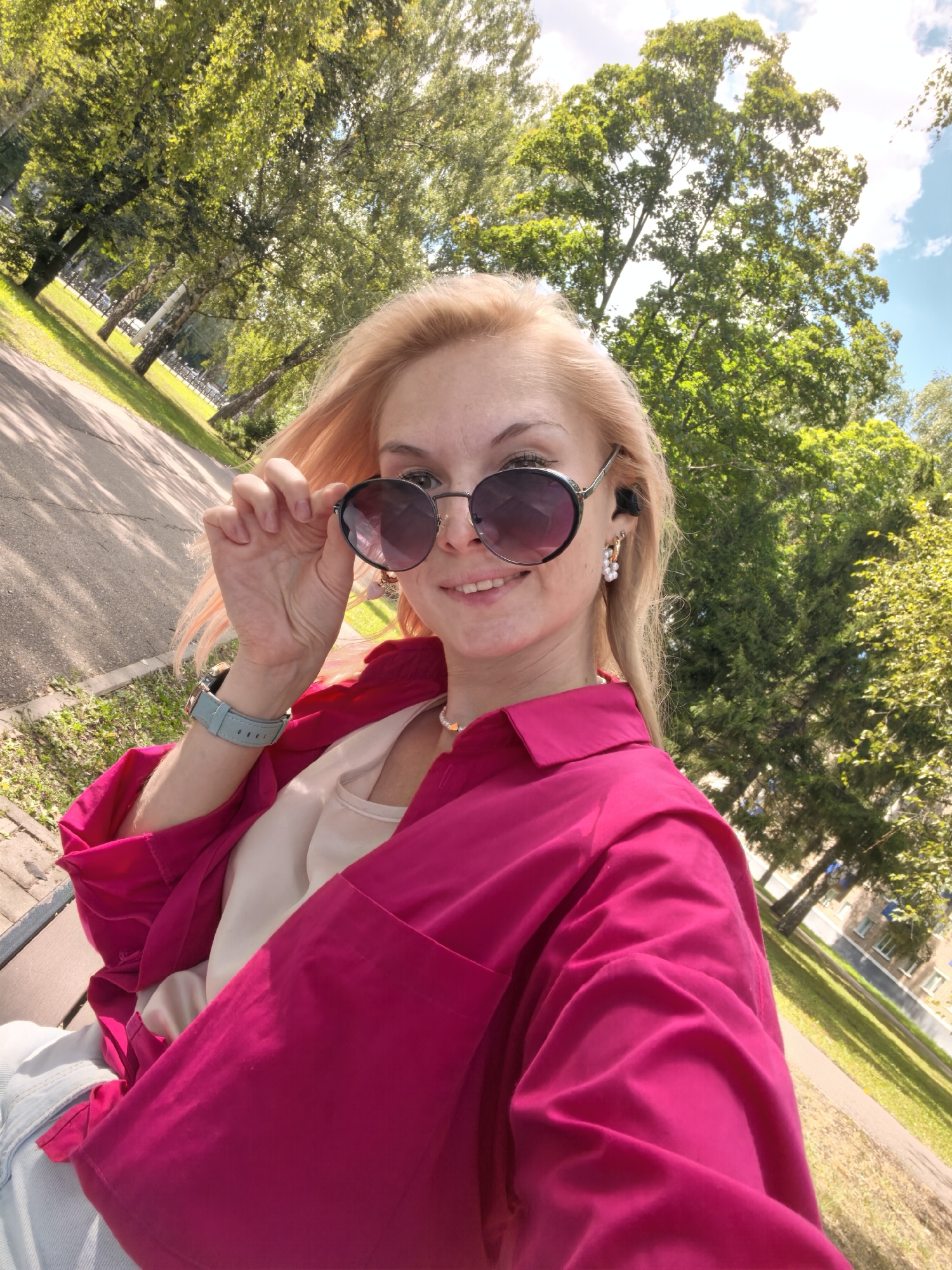 A woman with blonde hair and sunglasses is taking a selfie in a park. She is wearing a bright pink shirt and a light-colored top. She has pearl earrings and a watch on her left wrist. The background shows trees, a path, and buildings, suggesting an urban park environment. The scene is consistent with a location in Sterlitamak, Russia, on a sunny day.