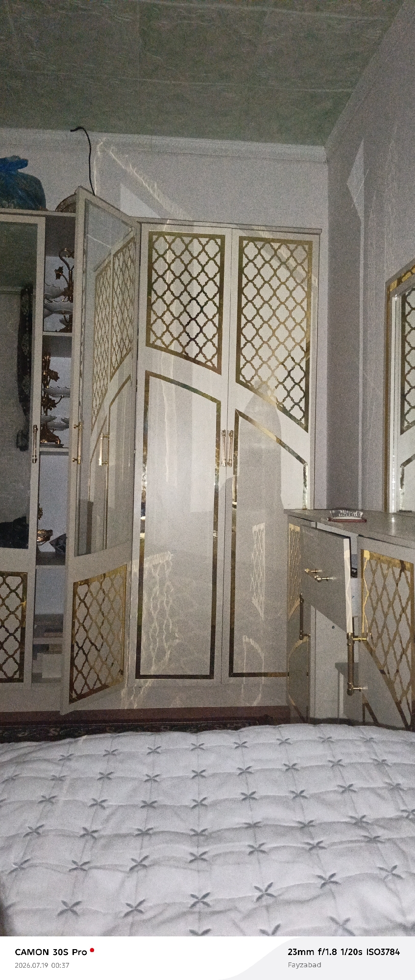 The image depicts an interior scene, likely a bedroom or dressing area, with a prominent wardrobe and a quilted bed in the foreground. The wardrobe is a tall, white structure with ornate gold detailing in a lattice pattern on its doors. It appears to be a built-in unit, with shelves visible in an open section on the left side, displaying decorative items. To the right of the wardrobe is a smaller cabinet or dresser, also with gold accents and an open drawer revealing its contents. The bed is covered with a white quilted comforter featuring a repeating cross-like pattern. The lighting suggests it is daytime, with some light reflecting off the wardrobe's surfaces. The camera information at the bottom indicates the photo was taken with a CAMON 30S Pro at 23mm focal length, f/1.8 aperture, 1/20s shutter speed, and ISO 3784. The location is identified as Fayzabad, Kalot, Tajikistan. No people are visible in the image.