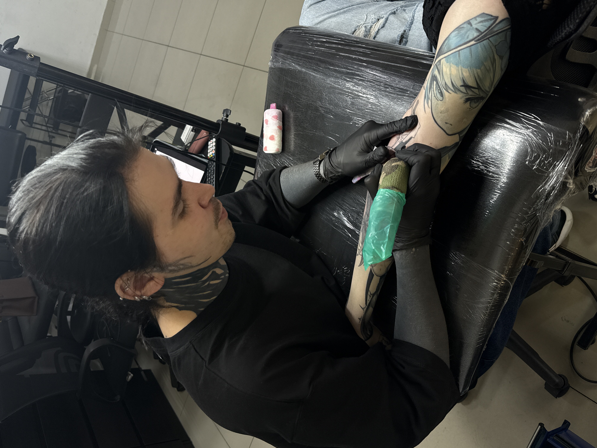 The image depicts a tattoo artist working on a client's forearm. The artist, a man with long dark hair streaked with grey, has extensive black tattoos covering his neck and part of his face. He wears black gloves and a black long-sleeved shirt. He is focused on the tattoo, holding a tattoo machine wrapped in a green protective sleeve. The client's arm is outstretched, and a tattoo of an anime-style female character with blue hair and eyes is visible on the upper part of the forearm. The client is sitting on a black tattoo chair covered in plastic wrap. In the background, a white bottle with a pink lid and small pink hearts is visible, along with a black metal stand and a tiled floor. The photograph was taken in Rostov-on-Don, Russia.