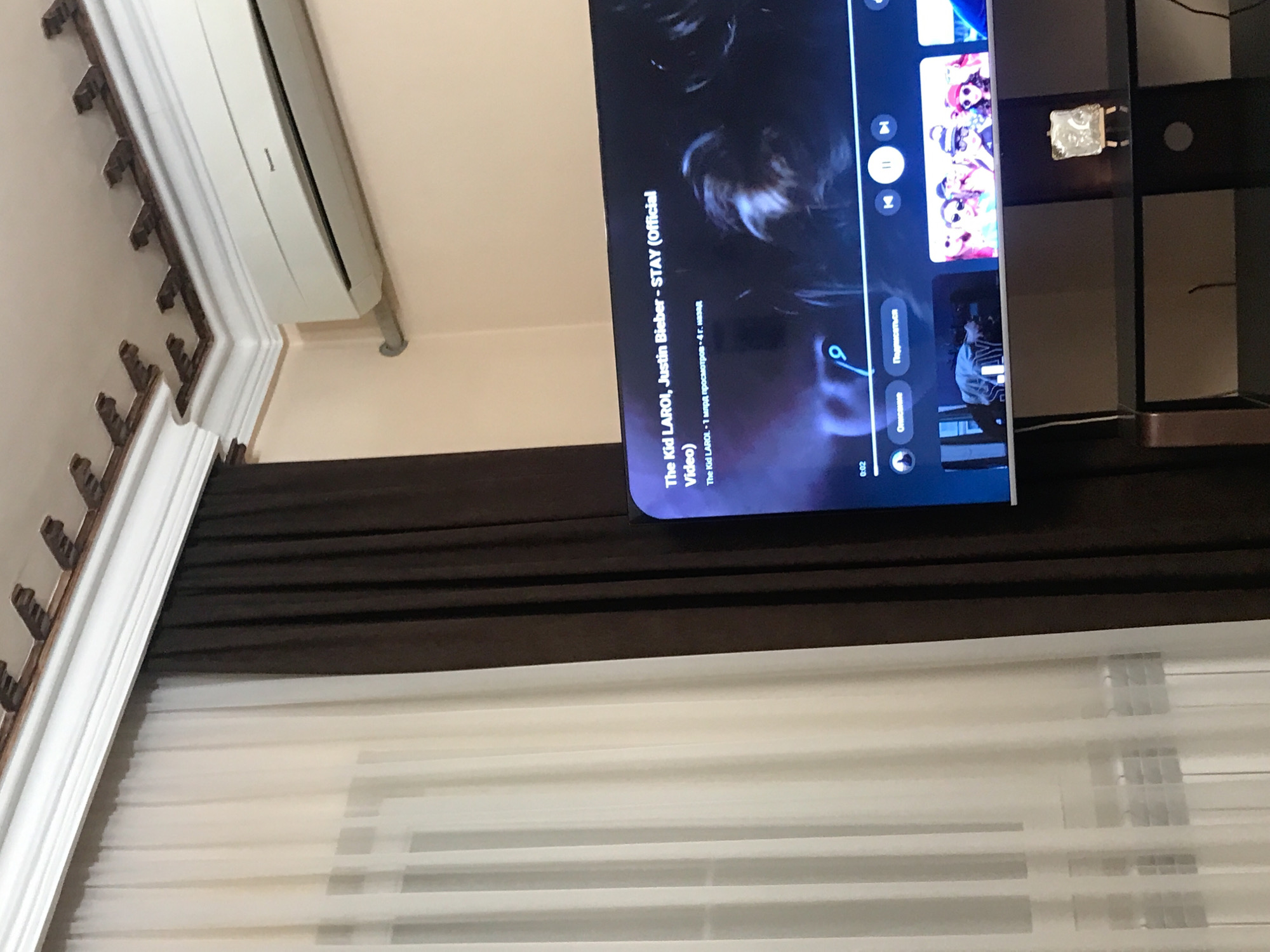 The image shows an indoor scene with a TV screen displaying a YouTube video of "STAY" by The Kid LAROI and Justin Bieber. Below the TV, there's a set of closed horizontal blinds. The interior features elements like decorative ceiling moldings and what appears to be an air conditioning unit. The location details, Yerevan, Armenia, are not visually confirmed in this image.