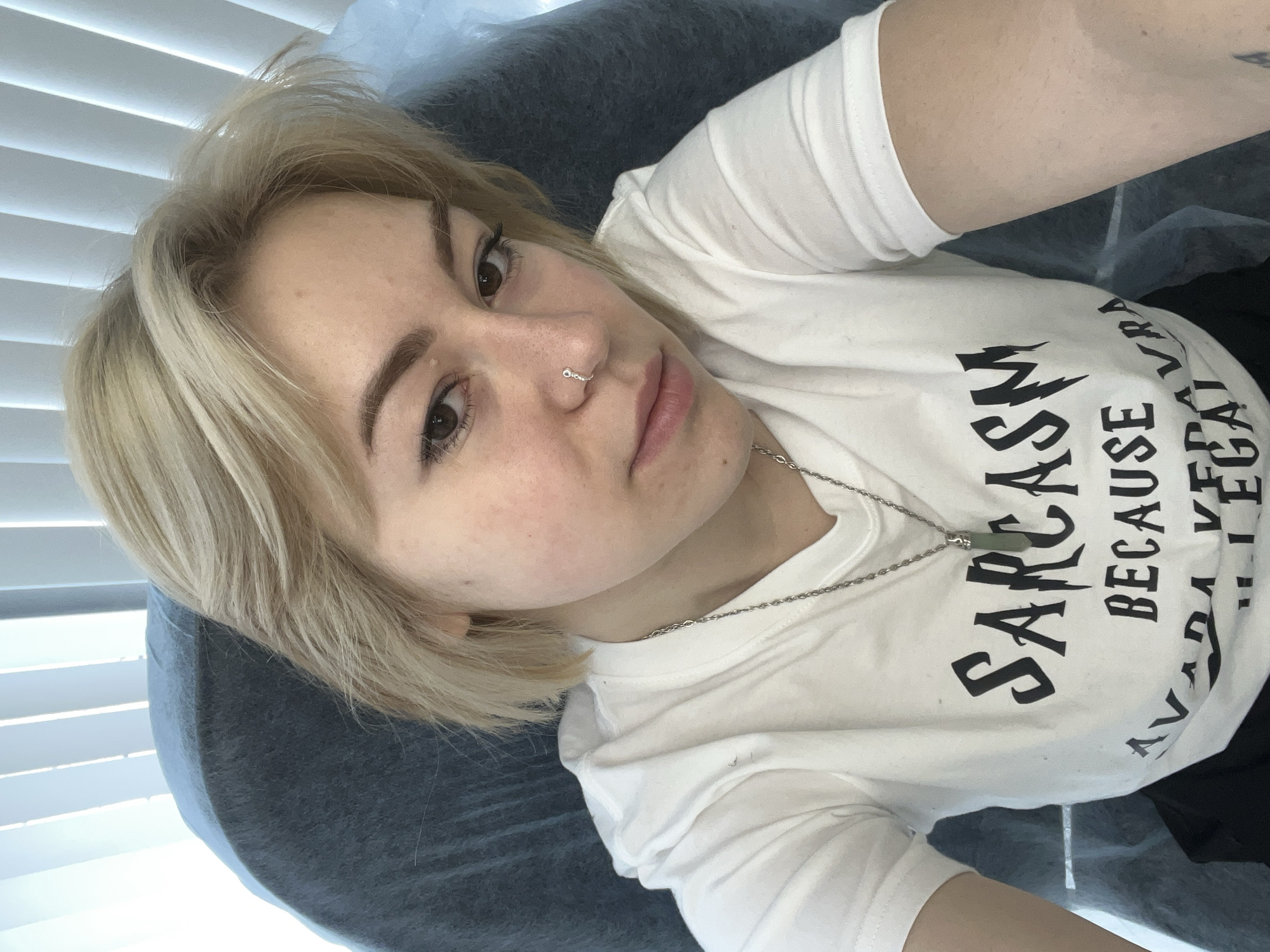 A young woman is captured in a close-up, eye-level selfie, lying down in an indoor setting in Odesa, Ukraine. She has short, light blonde hair that is slightly tousled around her face. Her complexion is fair with visible freckles, and she has dark, expressive eyes and neatly shaped eyebrows. A small, silver septum piercing adorns her nose.
She is wearing a white, short-sleeved t-shirt featuring bold black text. The most prominent words visible are "SARCASM" and "BECAUSE," with additional fragmented text like "AVAILA" suggesting a humorous phrase, possibly "SARCASM BECAUSE BEATING PEOPLE IS ILLEGAL." A silver chain necklace with a rectangular, pale green pendant rests on her shirt.
The woman's gaze is directed calmly at the camera, conveying a neutral expression. She is lying on a dark blue or grey upholstered surface, possibly an examination table or salon chair, which is partially covered with a light, disposable sheet. In the upper left background, white horizontal window blinds are visible, indicating natural daylight filling the room. The overall lighting is soft and even, suggesting it is daytime.