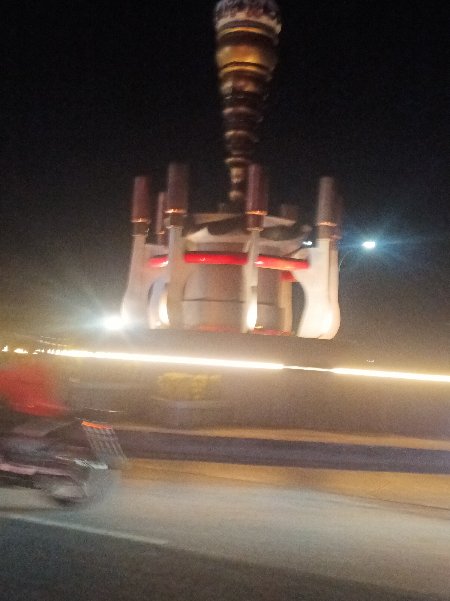 The image shows a large, metallic, industrial-looking object suspended from above, possibly a crane or part of a construction project. The object is cylindrical at the top and expands into a more complex, cage-like structure with red accents below. It is illuminated by artificial lights. The background suggests an outdoor, nighttime environment, possibly a roadway or industrial area, with blurred lights from vehicles and other structures. The lower portion of the image is highly blurred and shows what appears to be a moving vehicle, possibly a truck, with red taillights. The overall impression is one of movement and activity in an urban or industrial setting at night. The provided context that this scene is located in Bantarsoka, Indonesia, places it within that specific geographic location.