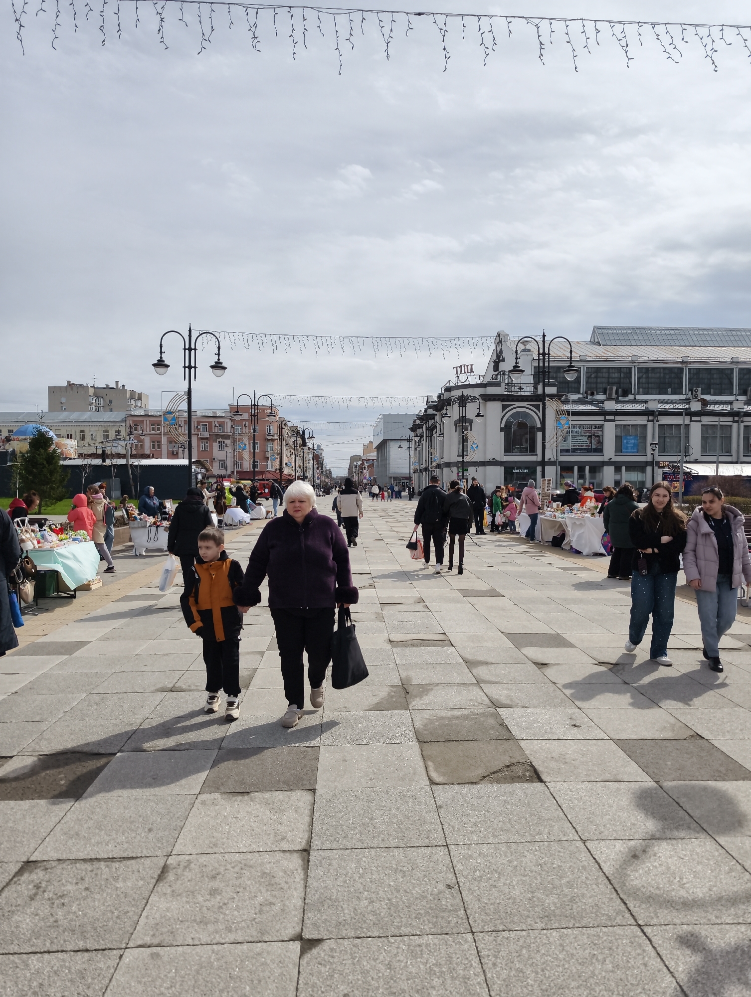 The image depicts a bustling city scene in Saratov, Russia. A woman and a child are walking hand-in-hand toward the viewer on a wide, paved pedestrian walkway. The walkway is composed of large square tiles in alternating shades of gray. Vendors have set up tables with merchandise along the sides of the walkway, suggesting a market or fair is taking place.
Other pedestrians are visible, walking in both directions along the walkway. Buildings line the street, displaying various architectural styles. Street lamps with ornate designs are positioned along the walkway. Decorative garlands are strung overhead. The sky is overcast, with a thin layer of clouds.