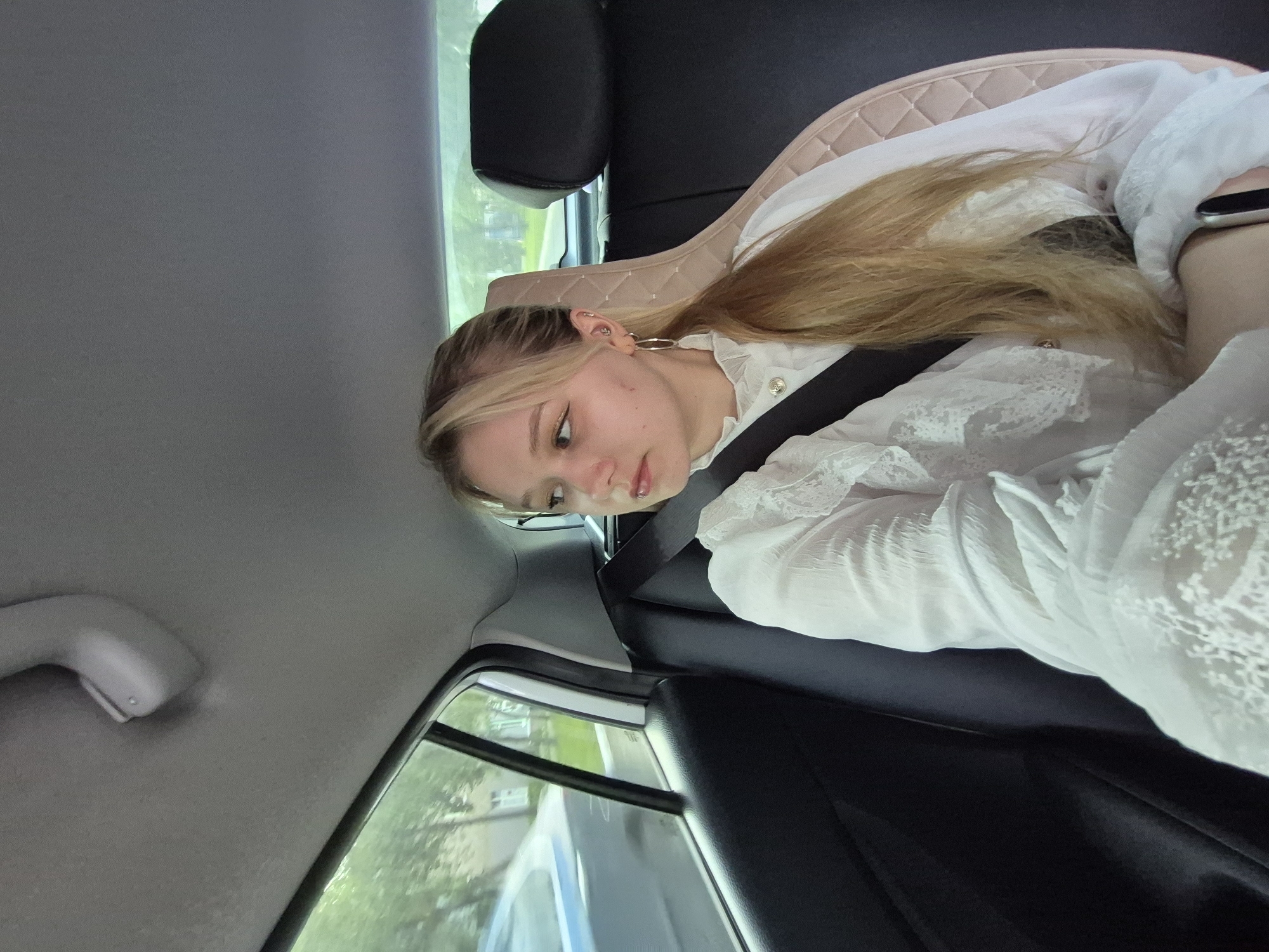 A young woman with blonde hair is seated in the back seat of a car, looking out the window. She is wearing a white, ruffled blouse and a seatbelt. Outside the car window, trees and buildings are visible, suggesting an urban or suburban environment. The interior of the car includes a dark-colored seat and a textured headrest. The scene appears to be set during the daytime, with natural light filtering into the car.