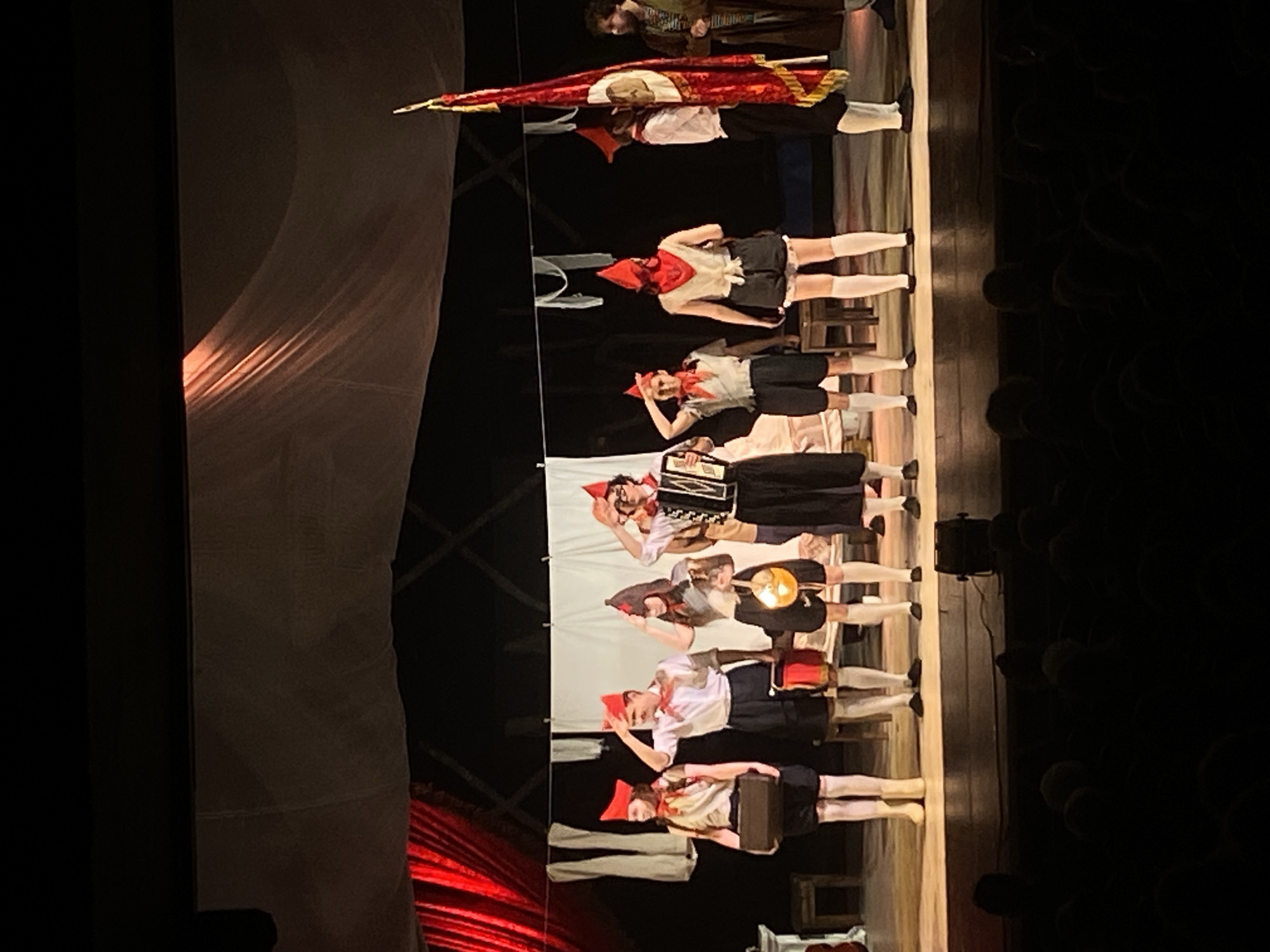 A stage performance in Unknown, Unknown, features a vertical line of actors on a stage. The performers are dressed in costumes that include red hats and scarves, white shirts, dark shorts or skirts, and white stockings. One performer is playing an accordion. Another is holding a lit lamp, while others hold props like suitcases and books. A red flag is held by the actors at the top of the line. Stage lighting illuminates the performers. A neutral colored backdrop hangs behind the actors, and the audience is visible in the dark area on the right. A reddish curtain hangs on the left side of the stage.