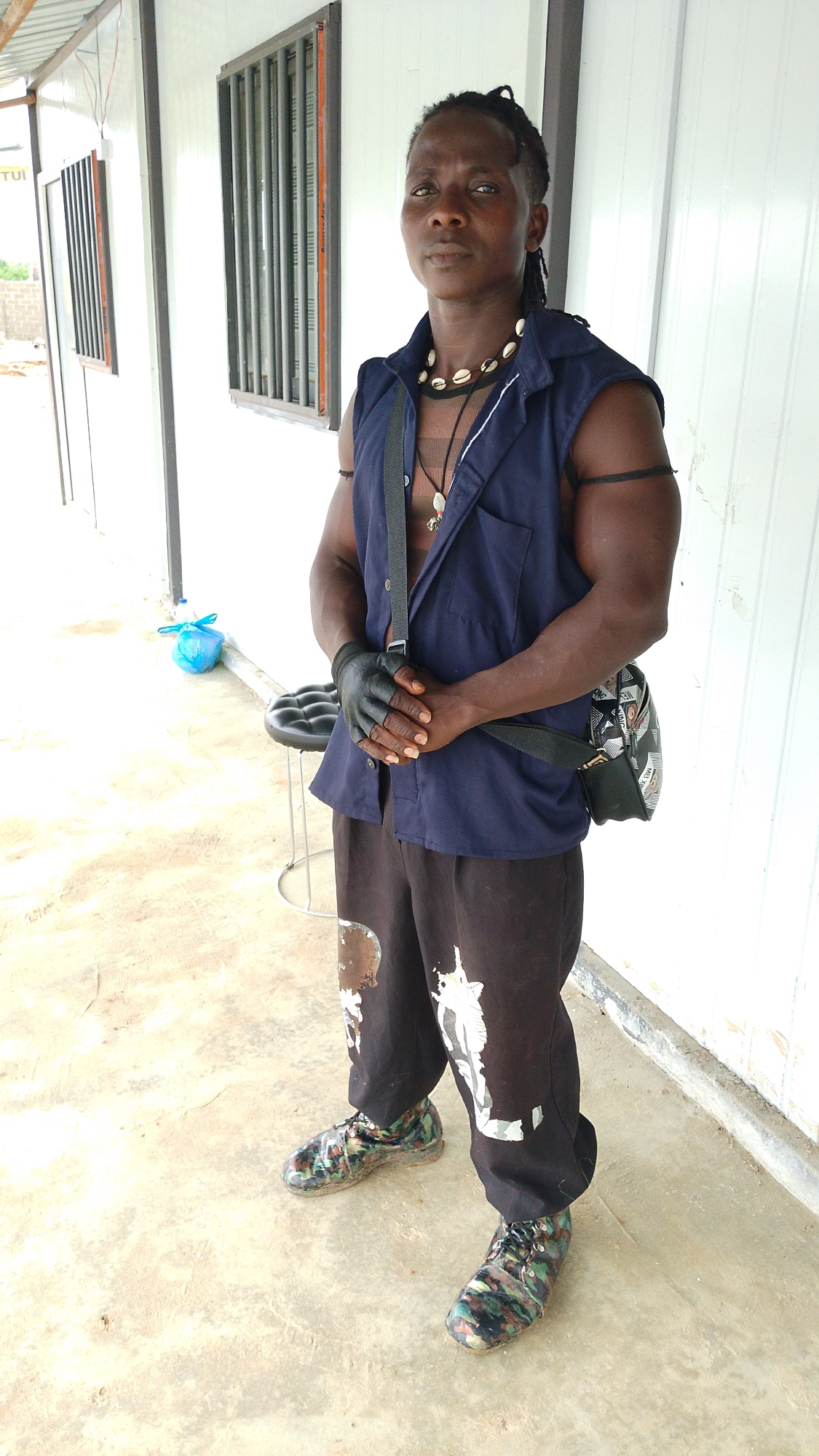 A Black male, appearing to be in his late 30s or early 40s, stands facing the camera on a concrete ground. He has dark skin, short dreadlocks, and is wearing a dark blue sleeveless vest over a black mesh shirt. He is also wearing black pants with white paint splatters and camouflage-patterned shoes. Around his neck, he wears a necklace with shell pendants. His left hand is wrapped in black tape. He has a dark-colored bag slung across his body. To his left, a blue plastic bag rests on the ground next to a small, round stool. Behind him, a white corrugated metal structure with windows is visible. The setting appears to be outdoors, possibly in a construction area, with a view of other structures and some vegetation in the background. The lighting suggests it is daytime. The image was taken at an elevated angle, looking down slightly at the subject.