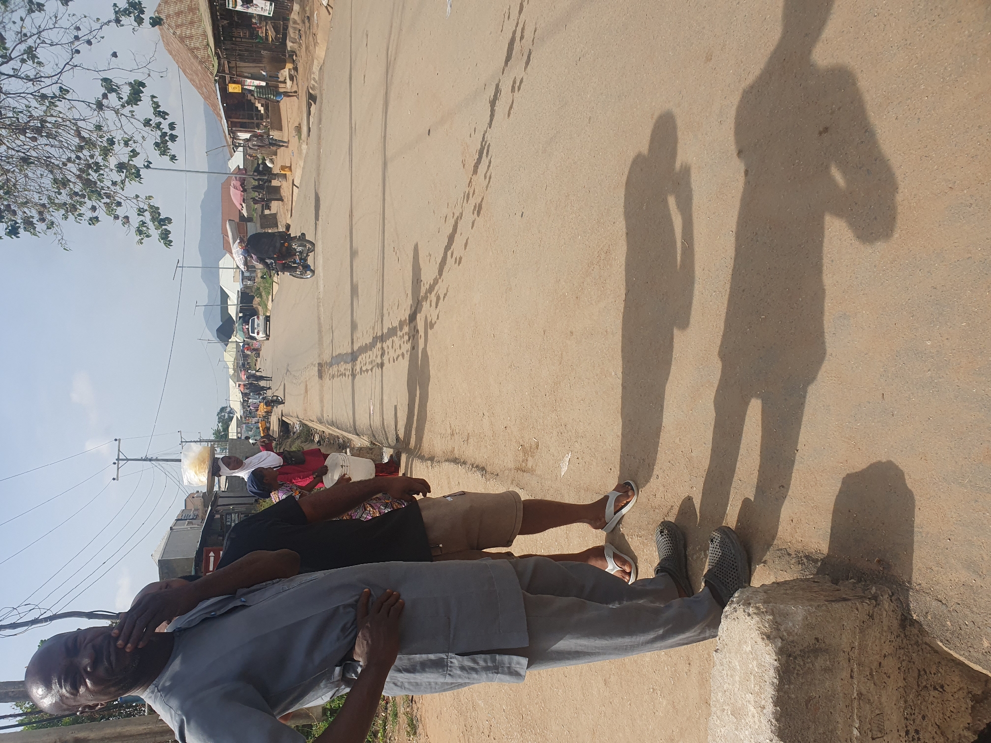 A daytime shot in Gwagwalada, Nigeria shows an outdoor scene along a street. The photo is oriented sideways.
Two people are visible near the bottom of the frame. One man wears a light gray outfit, while another wears a black shirt and khaki shorts. They stand near what appears to be a concrete curb.
Further down the street, people are carrying items on their heads, and a motorbike is visible. Shadows cast by people are seen on the ground. In the distance, buildings are discernible against a mountain backdrop under a bright sky. The street surface is rough and uneven. Trees and power lines are also present.