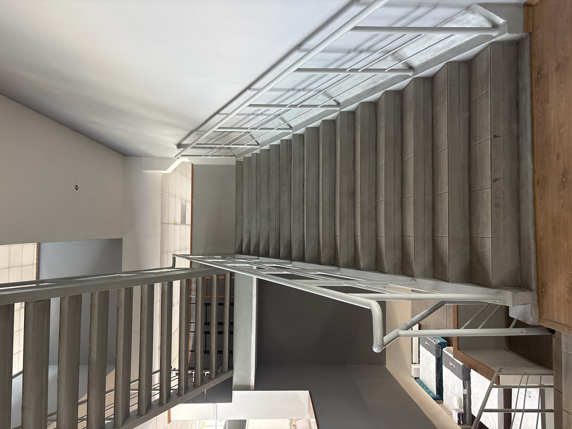 The image shows a view of an indoor staircase. The staircase has concrete steps and a white metal railing. The walls are painted white and gray. There is another metal staircase visible in the background. In the lower left corner, there are some mattresses displayed. The lighting suggests an indoor environment, possibly a commercial building or apartment complex. The image does not provide any specific information to determine its location in Zaporizhzhia, Ukraine.