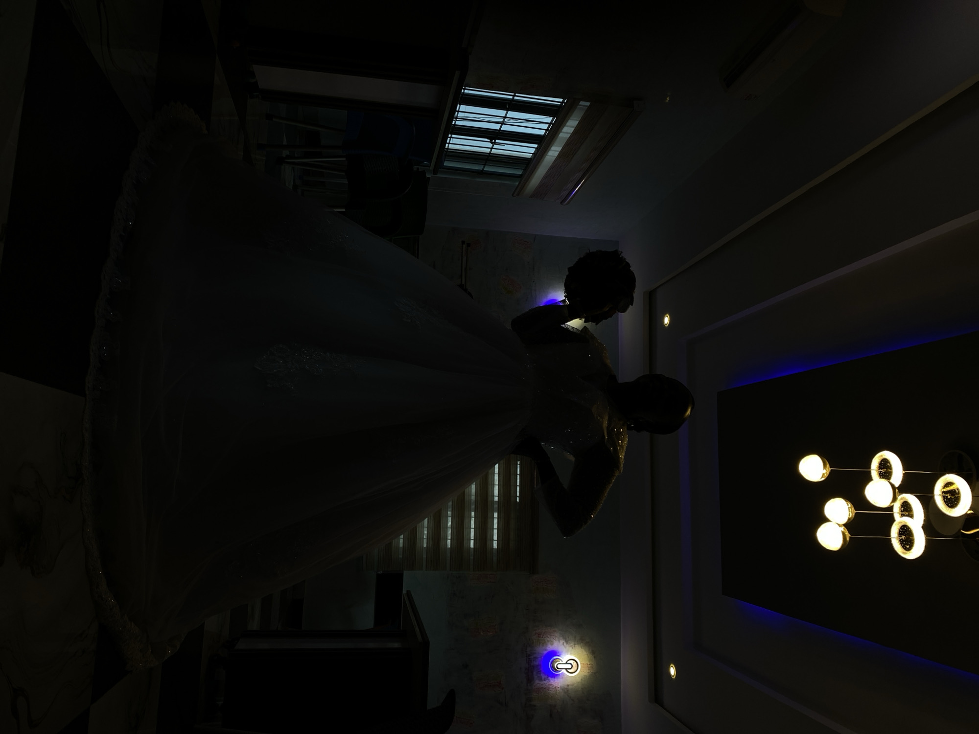 A person wearing a white gown is the central figure in a dimly lit interior space. The room features a window with visible blinds, a decorative ceiling light fixture, and subtle wall lighting. The location is potentially a room in Kuru Dungus, Nigeria. The orientation of the image is skewed.