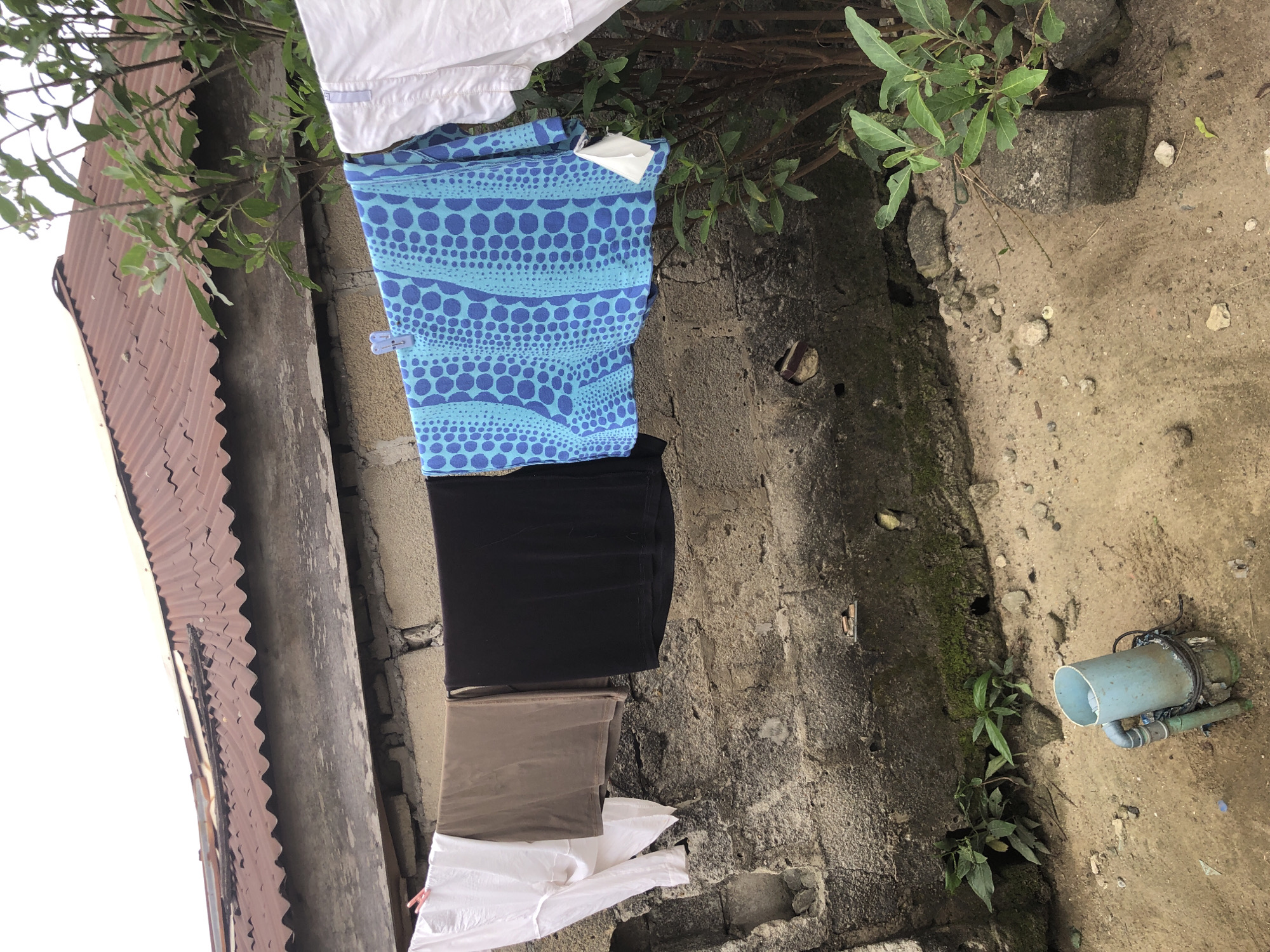 In the city of Ado, Nigeria, a clothesline hangs across a weathered concrete wall. On the line, several garments are drying: a white shirt, a brown t-shirt, a black t-shirt, and a blue and white patterned shirt. A small white cloth is also pinned to the blue and white shirt. Behind the clothesline, a corrugated metal roof is visible, and a tree with green leaves grows on the right side of the frame. In the foreground, on the sandy ground, a blue pipe with a green base sits on the dirt. The overall scene depicts an outdoor domestic setting.