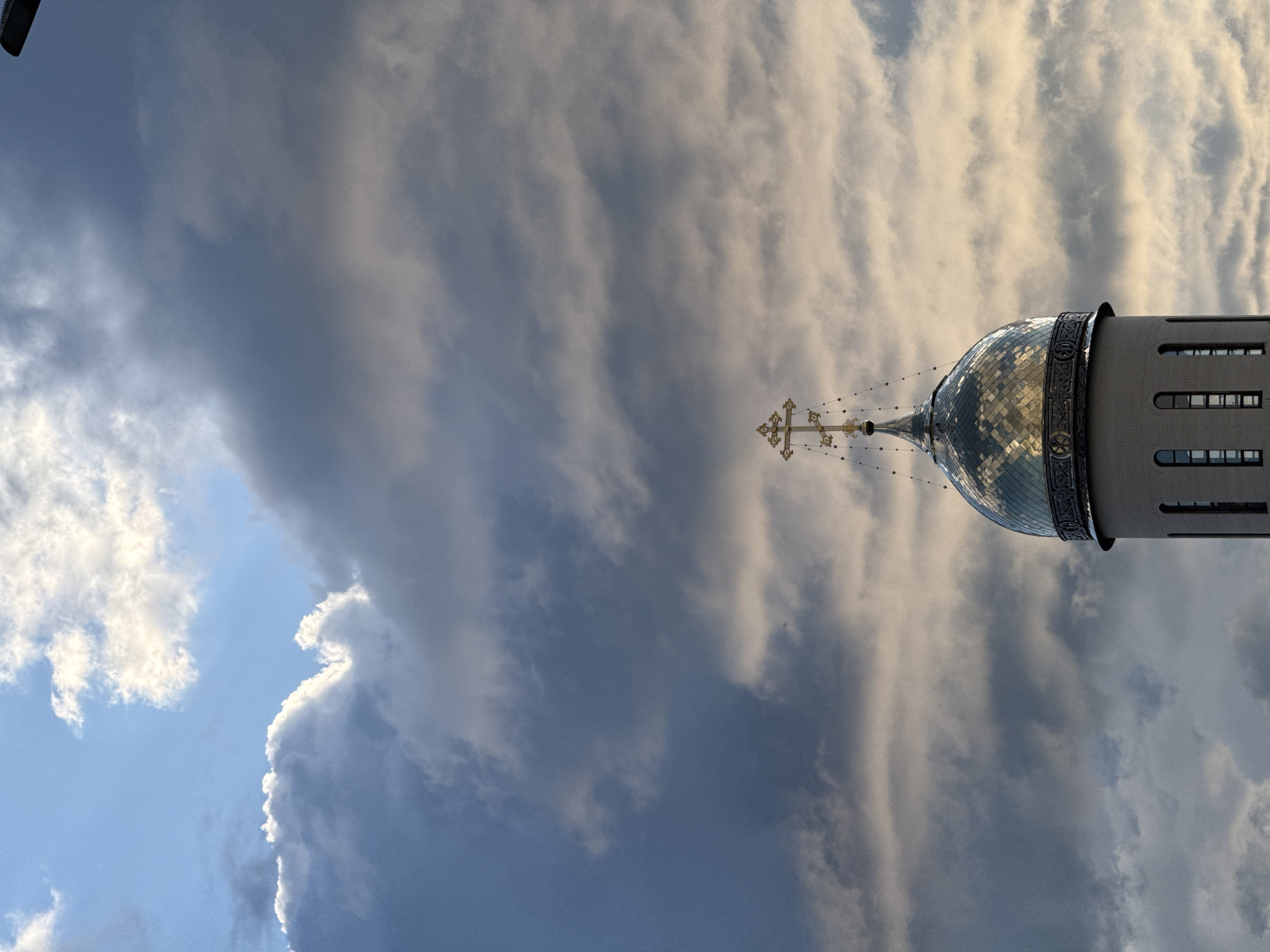 The image captures a close-up view of the dome and cross of a church in Saint Petersburg, Russia, against a dramatic sky filled with dark, textured clouds interspersed with patches of bright blue sky and sunlight. The dome is made of reflective material, likely metal, and the sunlight creates a mosaic of light and shadow across its surface. The cross atop the dome is ornate and golden, with a traditional Orthodox design. The church tower is constructed of light-colored brick and features a series of arched windows. The overall impression is one of religious architecture set against a dynamic and somewhat moody atmospheric backdrop. The specific location within Saint Petersburg is not identifiable from the image alone.