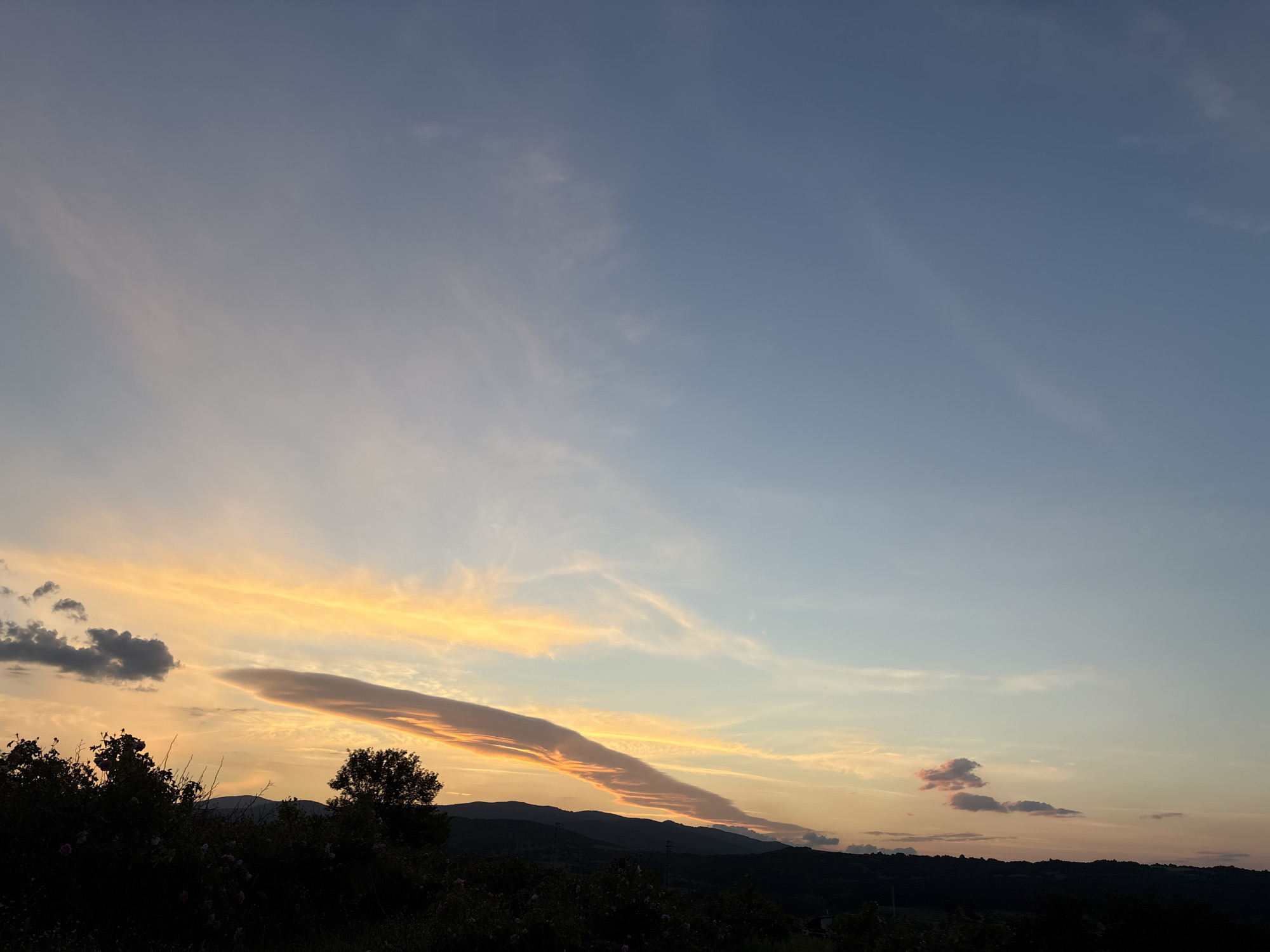 The image shows a landscape at sunset, with a dark silhouette of shrubs and trees in the foreground. Beyond the vegetation, there is a series of low hills or mountains. The sky is the focal point, with a gradient of colors ranging from deep blue in the upper right to orange and yellow near the horizon. A long, narrow cloud stretches across the sky, illuminated by the setting sun. Smaller, scattered clouds are also visible. There is no evidence this picture was taken in the city of Strelcha, Bulgaria.