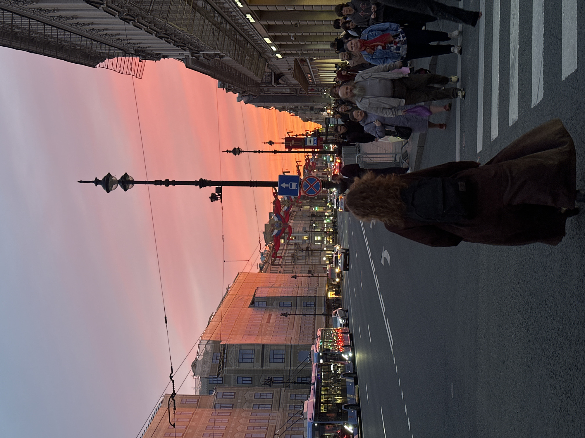 The image depicts a street scene in St. Petersburg, Russia, at sunset. Pedestrians are crossing a zebra crossing, and buses and cars are driving on the road. Buildings line both sides of the street. The sky is filled with pink and orange hues. A person with curly hair and a backpack is walking away from the camera. A streetlight is visible on the left side of the image, and traffic signs are visible in the center.