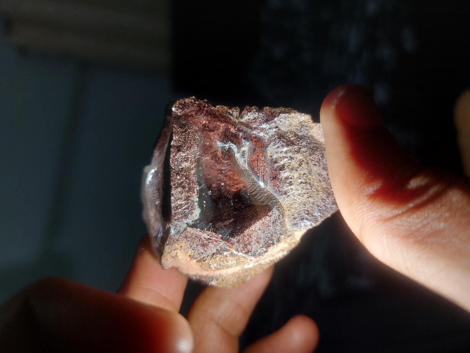 A person is holding a translucent, reddish-brown crystalline rock in their hand. The rock appears to have a rough, textured surface with some clear areas and a metallic sheen. The background is dark and blurry, suggesting a low-light environment. The presence of human fingers indicates the scale and context of the image. The location in Dar es-Salaam, Tanzania, is not evident from the image alone.