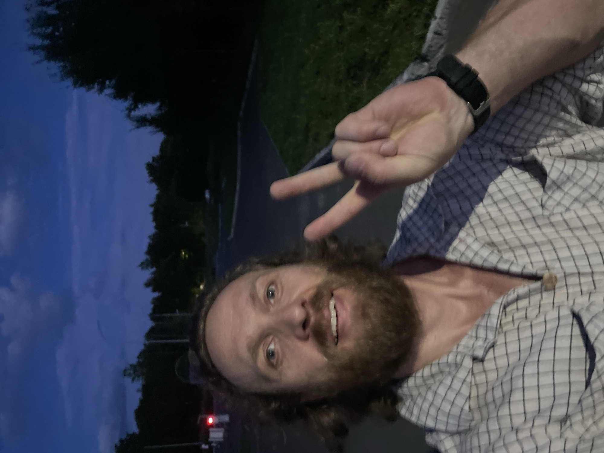 A man is taking a selfie outdoors at dusk. He has a beard and is wearing a plaid shirt and a black watch. He is holding up two fingers in a peace sign. The background shows trees and a cloudy sky. The location is identified as Kornevo, Russia.