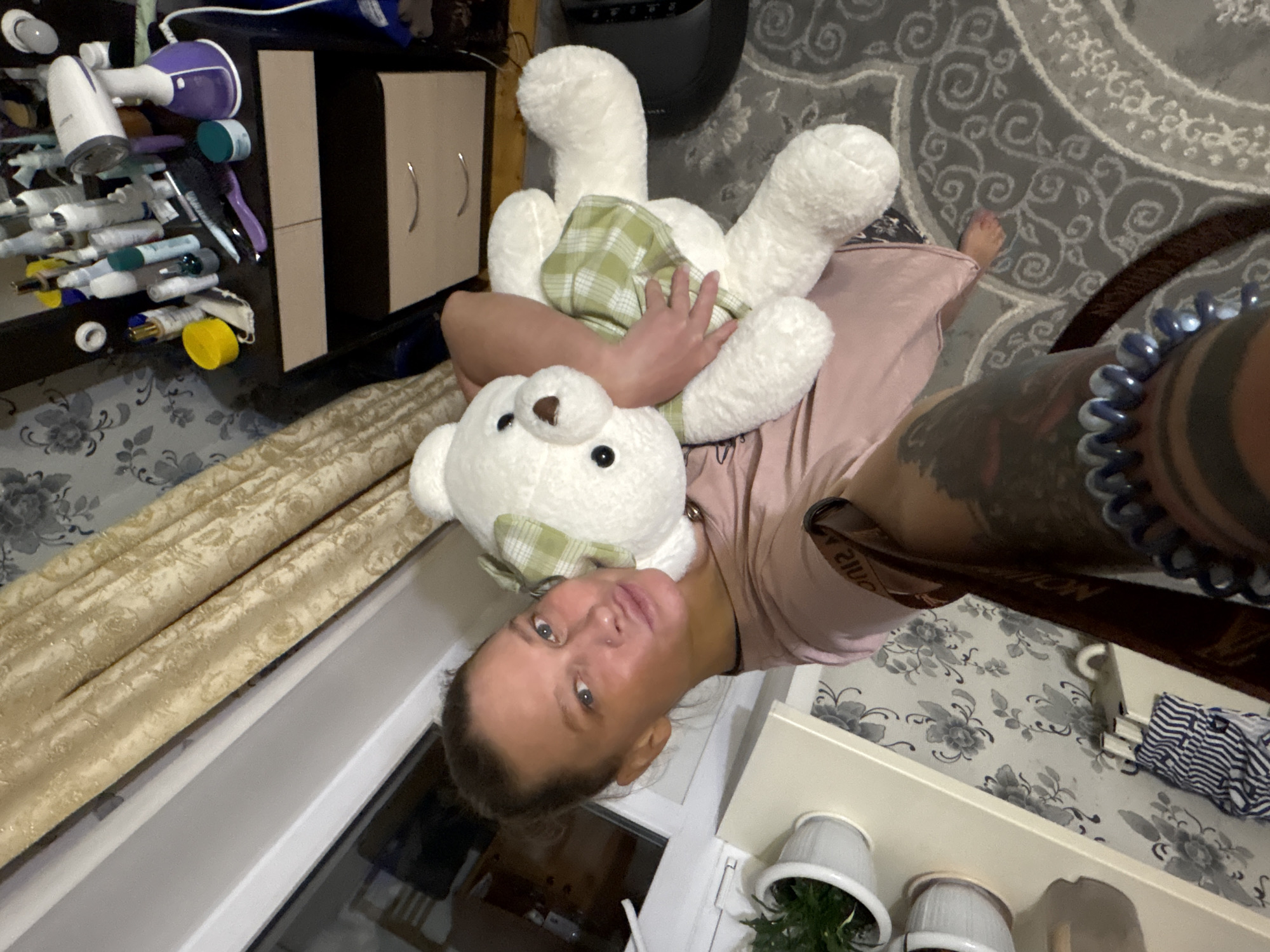 A person with tattoos on their right arm holds a large, white teddy bear with a green plaid bow and a matching vest. The person is in a room with floral wallpaper, a beige curtain, and a window. There are also potted plants on a shelf and various items on a dark-colored table, including a clothes steamer and toiletries. The carpet has a swirling pattern. While the location is not explicitly stated, it is claimed in the prompt to be Perm, Russia.