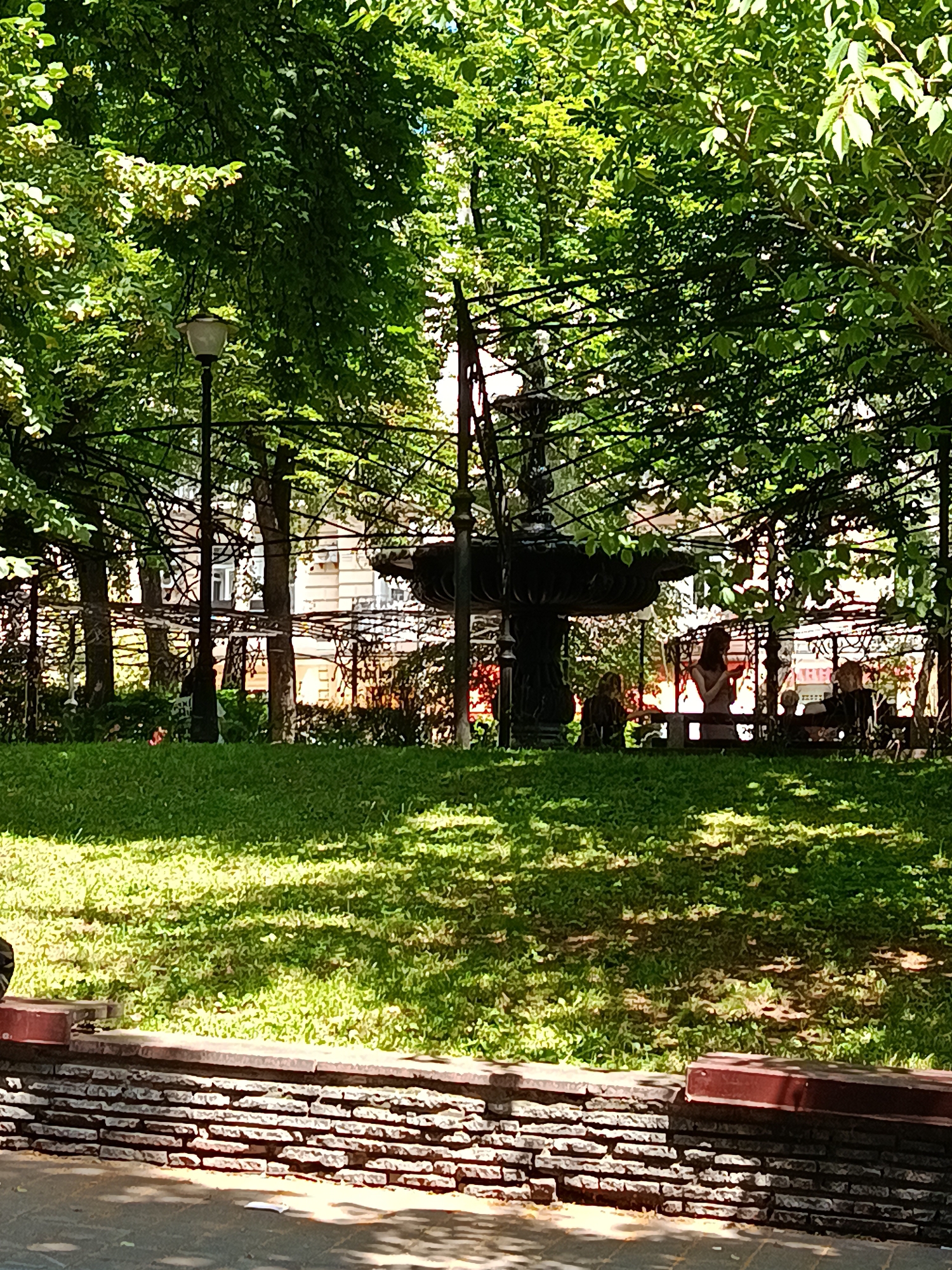 The image depicts a public park scene in Kyiv, Ukraine, likely on a sunny day with dappled sunlight filtering through dense green foliage. In the mid-ground, a tiered, dark-colored fountain stands as a central feature, surrounded by trees and a wrought-iron fence structure. Several individuals are seated or standing near the fountain and along benches in its vicinity, though their features are indistinct due to the distance and resolution. A well-maintained lawn occupies the foreground, bordered by a low, decorative brick wall topped with dark red seating elements. Beyond the park's greenery, multi-story buildings with light-colored facades are visible, suggesting an urban setting. A lamppost with a modern fixture is also present to the left of the fountain. The overall atmosphere is serene and typical of a city park.